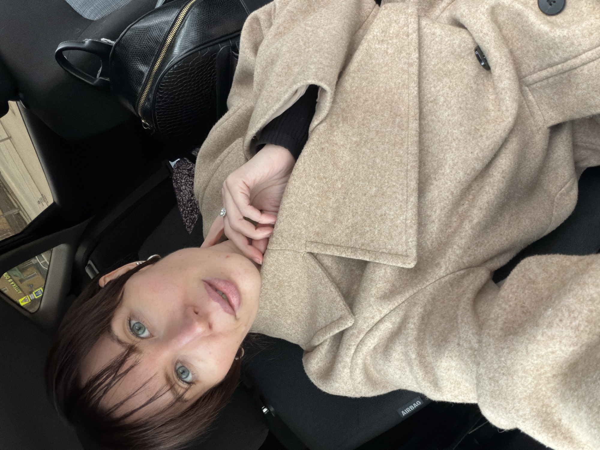 The image features a person, likely female, with shoulder-length dark brown hair and blue eyes, captured in a selfie while reclining in what appears to be the interior of a car. Her head is tilted, and she looks directly at the camera with a neutral expression. She is wearing a light beige or tan-colored coat, possibly made of wool, which has visible black buttons. A black long-sleeved top is visible underneath the coat.
One of her hands is near her face, revealing a ring with a clear stone on her finger. A black faux leather backpack with a gold zipper is positioned on an adjacent seat, partially visible on the left side of the frame. The car's interior features black seats and a panel with the word "AIRBAG" clearly printed in white text. In the reflection of the side mirror, located in the upper left corner, a blurred urban or street scene can be observed, along with reversed Cyrillic text, which is likely a street sign or advertisement from Perm, Russia. The lighting suggests it is daytime.