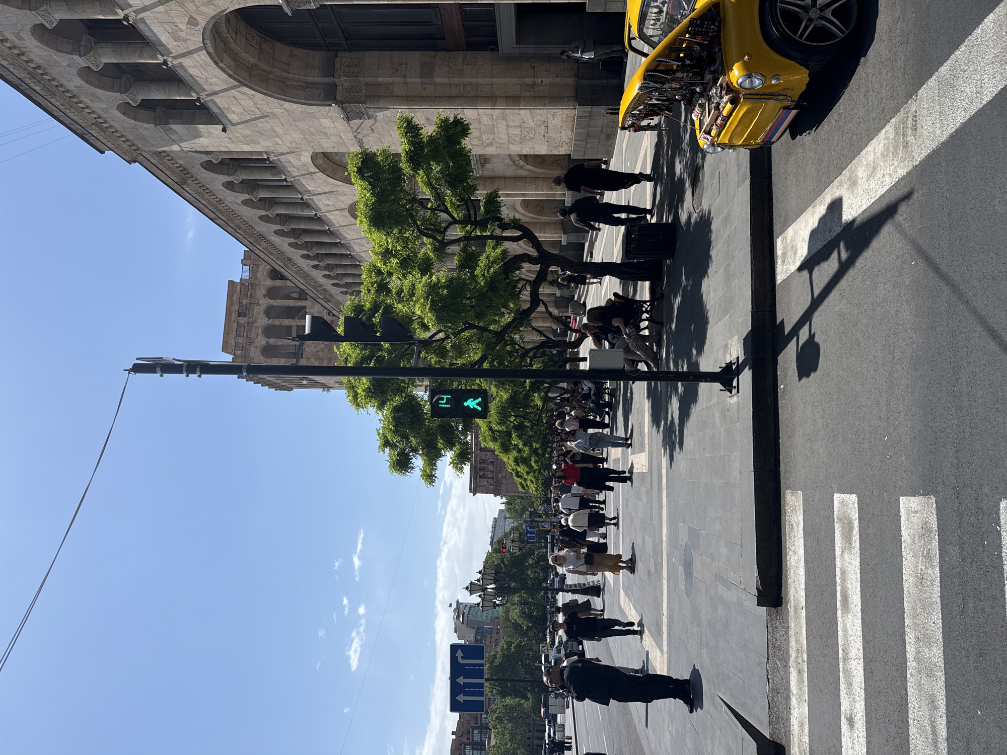 The image shows a city street in Yerevan, Armenia. A large building with arched architectural details lines the left side of the street. A yellow vintage car with the hood open is parked on the right.
A group of people are gathered on the sidewalk, near trees and benches. A traffic light shows a green walking signal. Further down the street, more pedestrians are visible. Traffic signs can be seen in the distance.
The sky is blue with scattered clouds. A crosswalk is visible in the foreground.