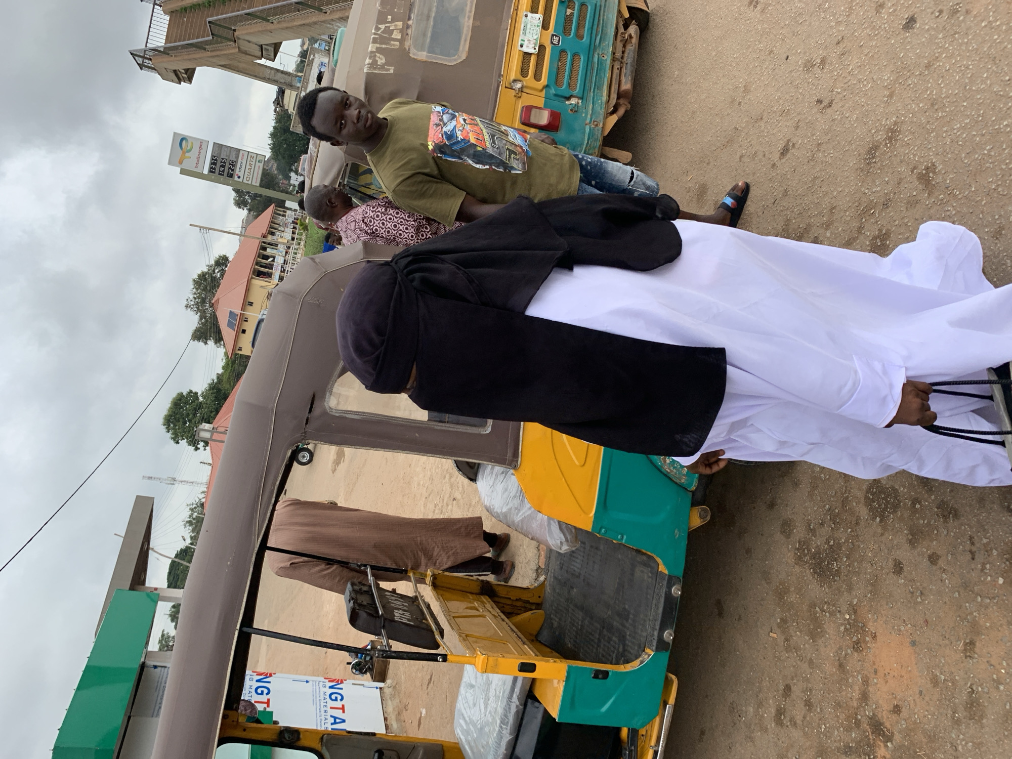 The photograph, taken in Gura Top, Nigeria, depicts an outdoor street scene on an overcast day, featuring several individuals and two tricycles, commonly known as "keke Napep." In the foreground, a person clad in white robes and a black head covering stands with their back to the camera, positioned next to a yellow and green tricycle. Adjacent to them, a man in a green t-shirt with a graphic design and ripped blue jeans leans against the tricycle, looking towards the camera. He wears black sandals. Partially visible behind him is another individual in patterned clothing, and a fourth person in brown attire is seen inside the tricycle.
The background features a prominent "TotalEnergies" fuel station sign displaying fuel prices: 177.5, 137.5, and 220, with "QUARTZ" also visible. Above the sign, a concrete pedestrian bridge spans the area. Beyond, buildings with orange-red roofs and green trees are visible under the cloudy sky, traversed by power lines. The tricycles bear text such as "NGTAI" and "IG MATERIALS." The ground is a worn asphalt or concrete surface. The scene captures a moment of everyday life, possibly at a roadside stop or a petrol station, with people gathered near the tricycles.