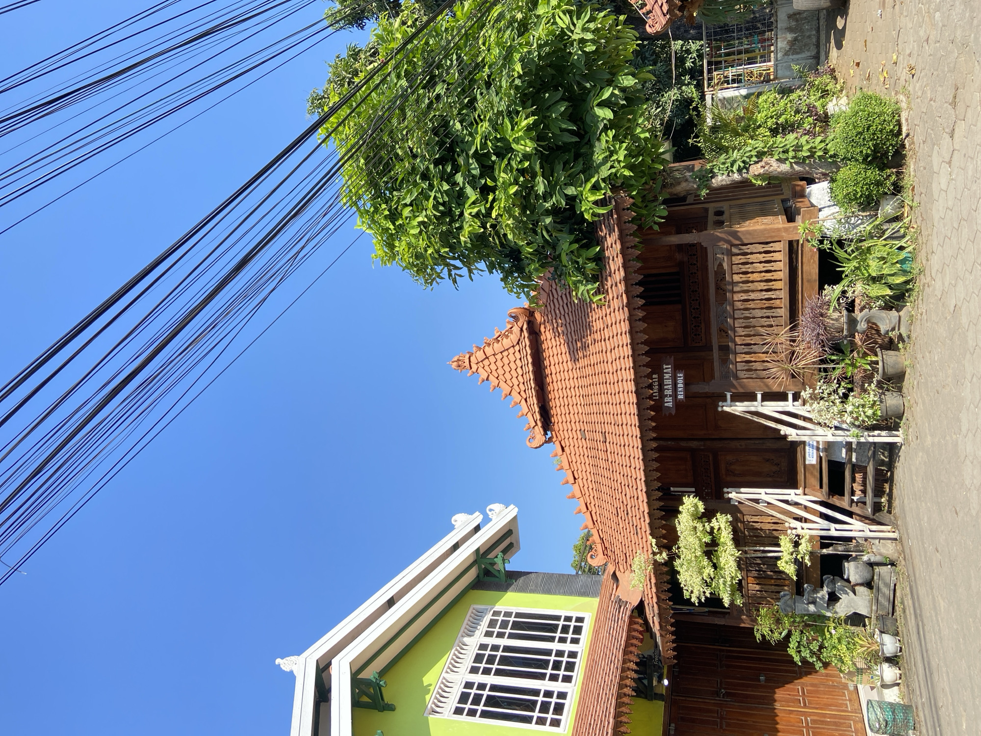 The image displays a building in Margorejo, Indonesia, with traditional Javanese architectural elements. A wooden structure with a terracotta tiled roof, featuring ornate carvings and a tiered design, stands prominently. A staircase with white railings leads up to the entrance of this structure. Adjacent to it, a lime green building with white trim and a large window with multiple panes is partially visible. The scene is set against a clear blue sky, with a dense green tree to the right of the wooden building. Overhead, a tangle of black electrical wires stretches across the frame. Several potted plants and decorative statues are arranged around the entrance of the wooden building. A sign above the entrance reads "LANGGAR AR-RAHMAT RENDOLE". The ground in front of the buildings is paved with interlocking concrete blocks. The image appears to be taken from a low angle, looking upwards.