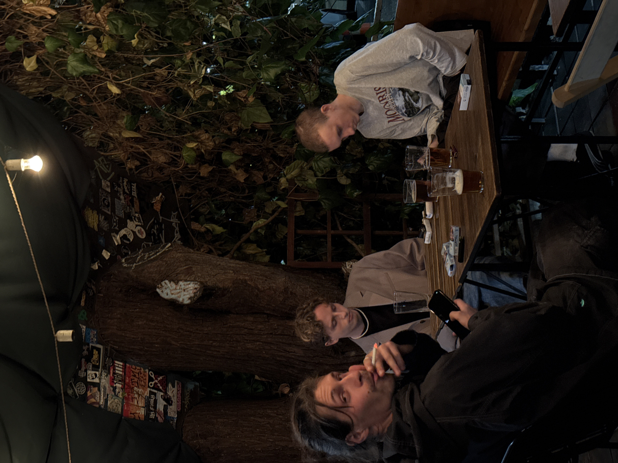 Three people sit around a wooden table outside. The table has several glasses, some filled with beer. One person in a light grey sweatshirt is looking down at the table, while another person in a light brown jacket is seated to his right. The third person is wearing a dark jacket. He has a cigarette in his mouth and is holding a mobile phone in his right hand. The area is decorated with vines and a tree. The location is Sochi, Russia.