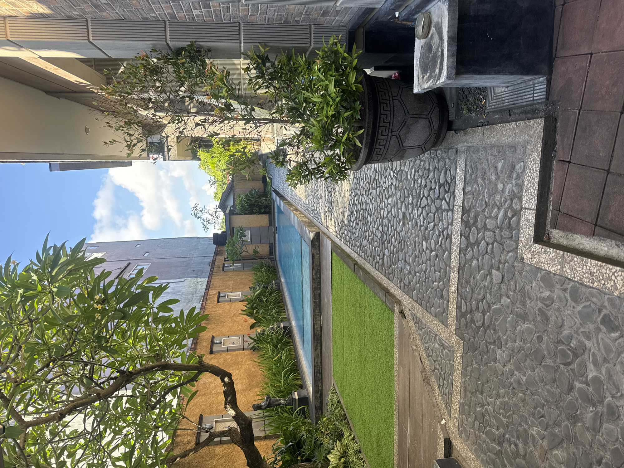 The image depicts a bright, outdoor scene, likely a private residence or resort area, characterized by a long, narrow layout featuring a path and a swimming pool, consistent with the tropical urban environment of Kuta, Indonesia. The perspective is taken from slightly above, looking down the length of the area.
The primary subject is a meticulously designed outdoor space. A textured path made of various grey pebbles set in concrete runs along the right side of the frame, with sections bordered by smaller, lighter aggregate. To the left of the path is a long, rectangular swimming pool with clear blue water. Adjacent to the pool, a strip of vibrant green artificial turf adds a contrasting element. Lush green plants and shrubs line the far side of the pool.
In the foreground, a large, ornate grey planter pot holds a leafy green plant. Further into the scene, a large tree with light green foliage and some visible white flowers (resembling plumeria) stands prominently, partially obscuring a building.
The setting includes various architectural elements. On the left, a multi-story building with a grey roof and some windows is visible under a clear blue sky dotted with fluffy white clouds. To the right of the pool, another building features an distinctive orange/yellow textured wall with several rectangular decorative accents that resemble small, recessed windows. Behind this, another building section appears to have a dark brick pattern.
The scene is devoid of people or activity, conveying a sense of serenity and stillness. The bright sunlight, indicated by the clear sky and distinct shadows, suggests it is daytime, likely late morning or early afternoon. No visible text is present in the image. The overall atmosphere is one of a well-maintained, tranquil outdoor space.