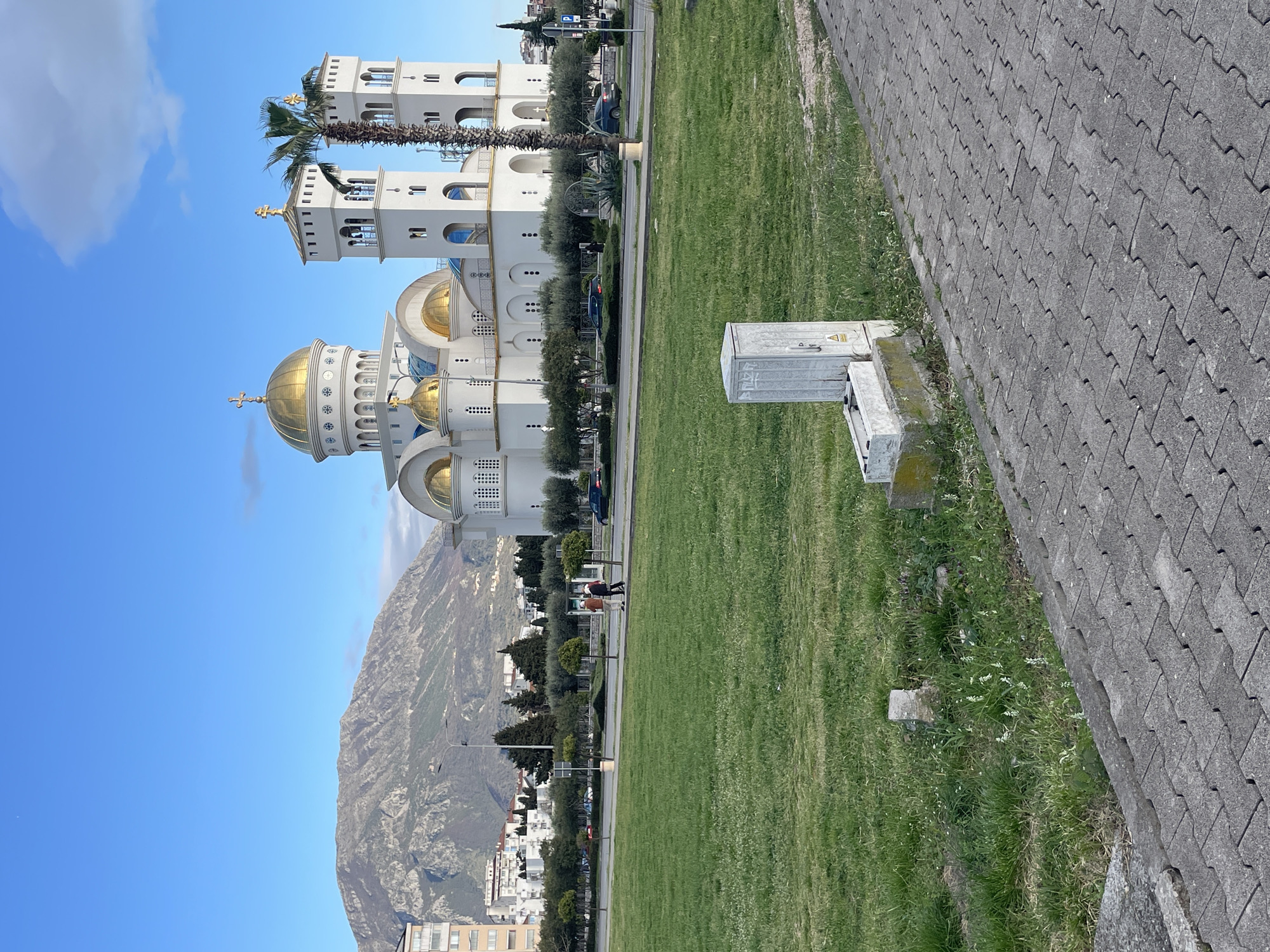 The image captures a view of the St. Jovan Vladimir Orthodox Church, a prominent landmark in Bar, Montenegro. The church's architecture features a large golden dome and two white towers, set against a clear blue sky. A mountain range provides a backdrop to the church and the city.
In the foreground, there is a green lawn that transitions to a gray brick walkway. A gray electrical utility box stands on the edge of the walkway. Several people are walking along the road adjacent to the lawn, some pushing strollers. Olive trees are visible along the road separating the church from the city.