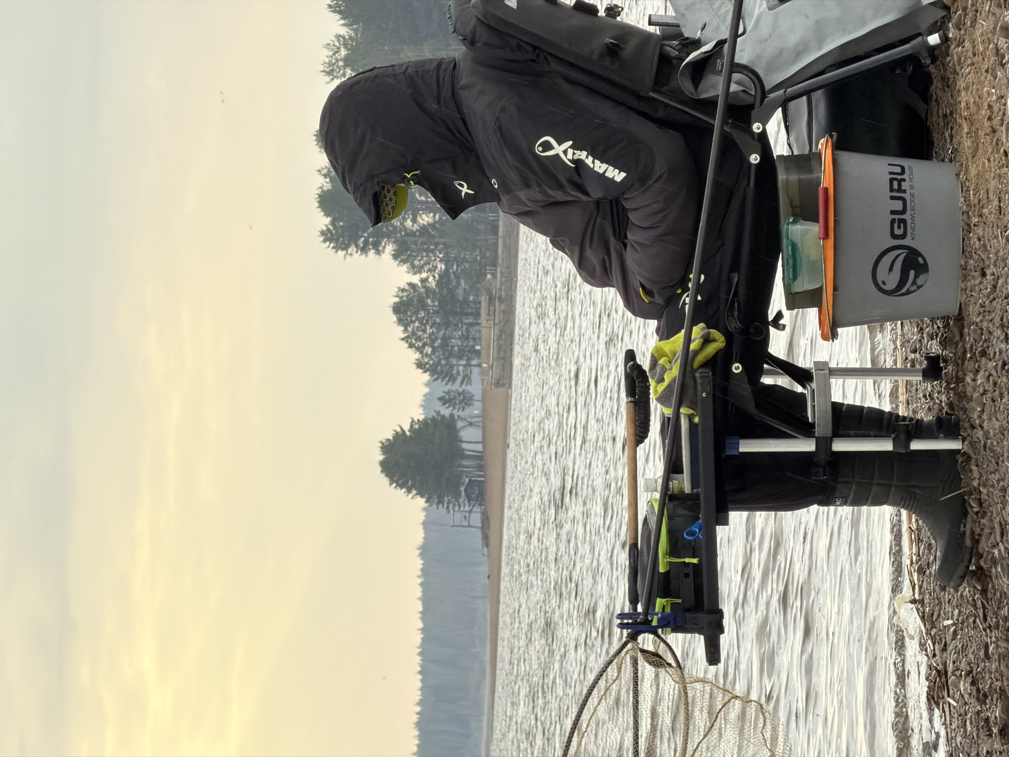 The image depicts a person engaged in fishing on the bank of a large body of water, likely a lake or wide river, in Chaikovsky, Russia. The individual is positioned on the right side of the frame, seated comfortably in a specialized fishing chair with various tackle and equipment arranged around them. They are dressed in a black, hooded, padded jacket featuring a white "MATRIX" logo with a stylized fish emblem on the chest and sleeve. Dark boots cover their feet, extending into the water's edge.
Beside the chair, a grey bucket with an orange rim prominently displays the brand "GURU KNOWLEDGE IS POWER" along with a fish-and-wave logo, containing smaller containers for bait or tackle. A fishing net is visible on the ground in the foreground, close to the water. The water itself is calm, showing slight ripples, and its surface is a muted grey, reflecting the overcast sky.
The scene is set on a cloudy day, with the sky appearing a soft, diffused yellow-grey, suggesting either early morning or late afternoon light. In the distance, across the wide expanse of water, a distinct shoreline is visible, densely covered with coniferous trees, possibly pines, and a few indistinct structures. The immediate ground where the person is seated is a mix of gravel and earth. Small, faint specks, possibly birds, can be seen in the sky. The overall impression is one of peaceful contemplation and focused activity in an outdoor natural setting.