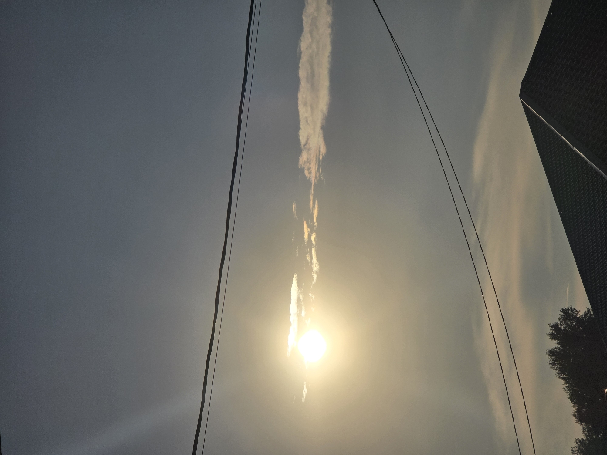 The image shows a sky with the sun visible, partially obscured by a thin, horizontal cloud. Two power lines run across the frame, one above and one below the sun. In the lower part of the image, the dark, peaked roof of a building is visible, along with the silhouettes of trees to the left. The scene appears to be set in the city of Ермолаево, Russia, possibly during a sunset or sunrise given the lighting. There are no people visible in the image. The overall environment is outdoors, under a cloudy sky.