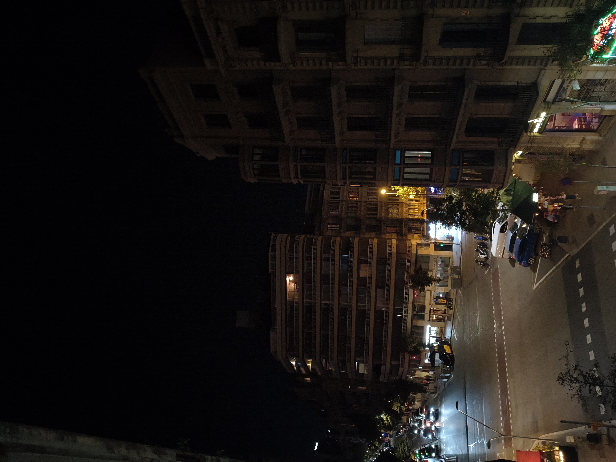 This is a nighttime street-level shot of a busy intersection in the Eixample district of Barcelona, Spain. Tall, multi-story buildings line the streets, illuminated by streetlights and the lights from within the buildings. The architecture is characteristic of Barcelona, with ornate balconies and repeating window patterns.
On the street, several cars are visible, including a yellow taxi, a blue car, and a white van. There are also a number of motorbikes and scooters parked and in motion. Pedestrians are walking on the sidewalks, some grouped together and others walking individually. One group appears to be seated at an outdoor cafe under a green awning.
The intersection is marked with traffic lights and crosswalks. The overall impression is of a vibrant urban environment at night, with the constant flow of traffic and people. The dark sky suggests it is late in the evening or early morning. The image captures the energy of city life.