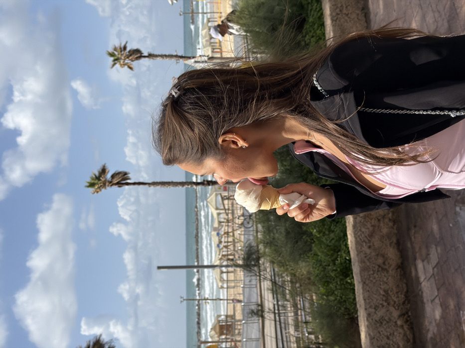 An image captured in Tel-Aviv, Israel, depicts a young adult female in profile, facing left, actively licking a light-colored ice cream cone held in her right hand. She wears a light pink top beneath an unzipped black jacket, with her medium-brown hair pulled back by a small, light-colored claw clip, revealing a stud earring in her right ear. She stands on a paved surface adjacent to a low stone wall covered with green shrubbery.
Beyond the immediate foreground, a street or promenade features a metal barrier or rail running parallel to a sandy beach. The beach is furnished with numerous light-colored sun shelters. The Mediterranean Sea, characterized by breaking waves and a visible breakwater in the distance, forms the horizon. Several tall palm trees are interspersed along the coastline, silhouetted against a partially cloudy blue sky. A blurred individual wearing a light-colored cap is visible on the beach in the far right background, and a bicycle is partially discernible behind the shrubbery.