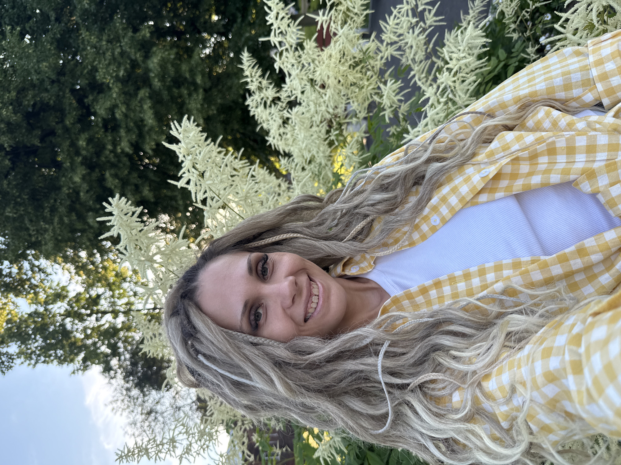 A woman with long, blonde, braided hair and a yellow and white checkered shirt is taking a selfie outdoors. She is smiling and looking directly at the camera. She is wearing a white tank top under her shirt. Behind her, there are feathery white flowers and green trees. The sky is visible in the upper left corner of the image. The location is not specified in the image.