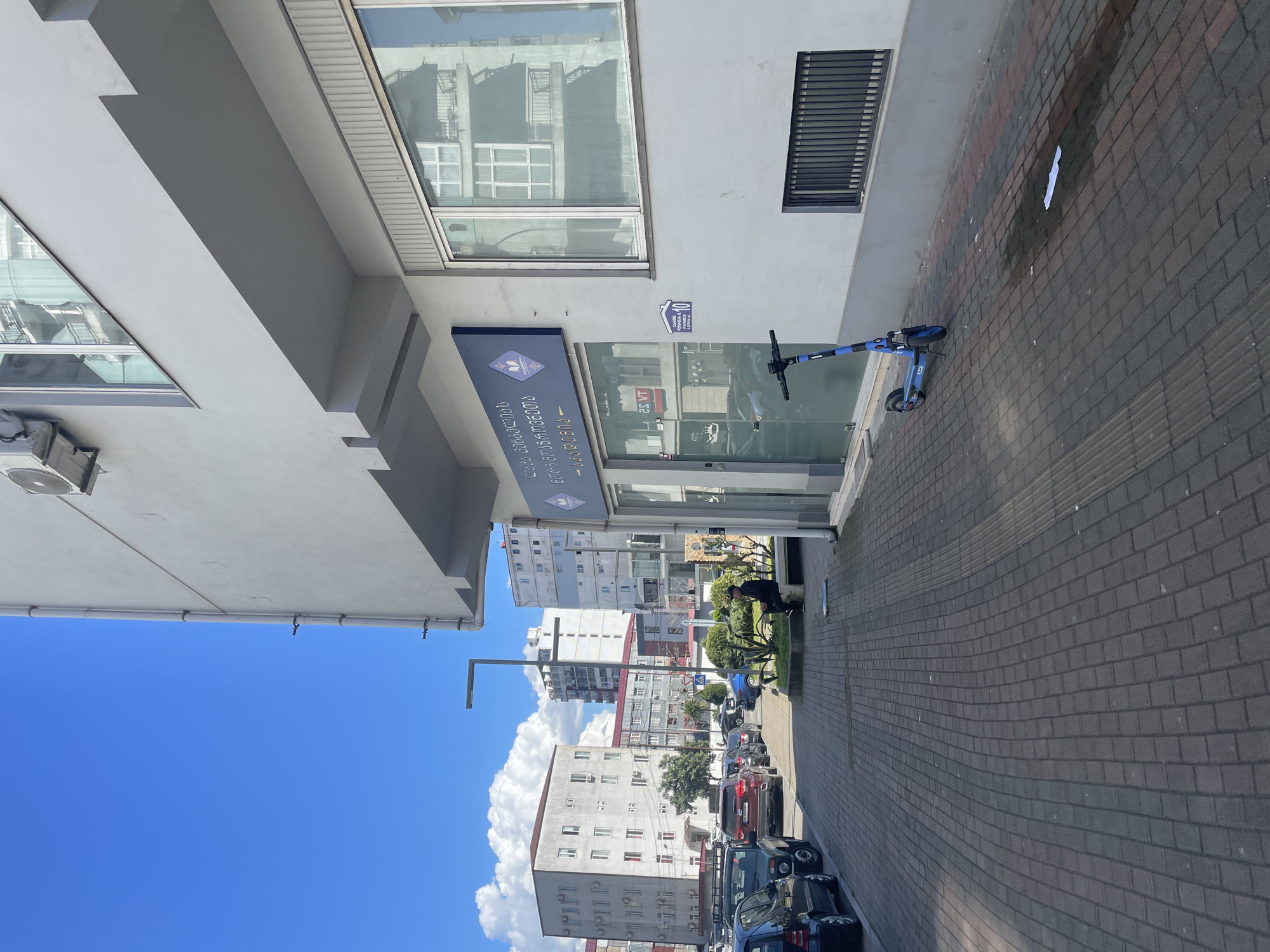 The image depicts a street scene in Batumi, Georgia. The sky is bright blue with scattered white clouds. On the left side of the image, a portion of a light-colored building is visible, with multiple window reflections. A sign with Georgian text and a logo is attached to the building's facade. A blue electric scooter is parked on the paved sidewalk in front of the building. The sidewalk is made of gray-red bricks. Further down the street, there are several multi-story buildings, with cars parked along the roadside. The buildings are mostly light-colored with multiple windows. Street lamps are visible along the sidewalk.