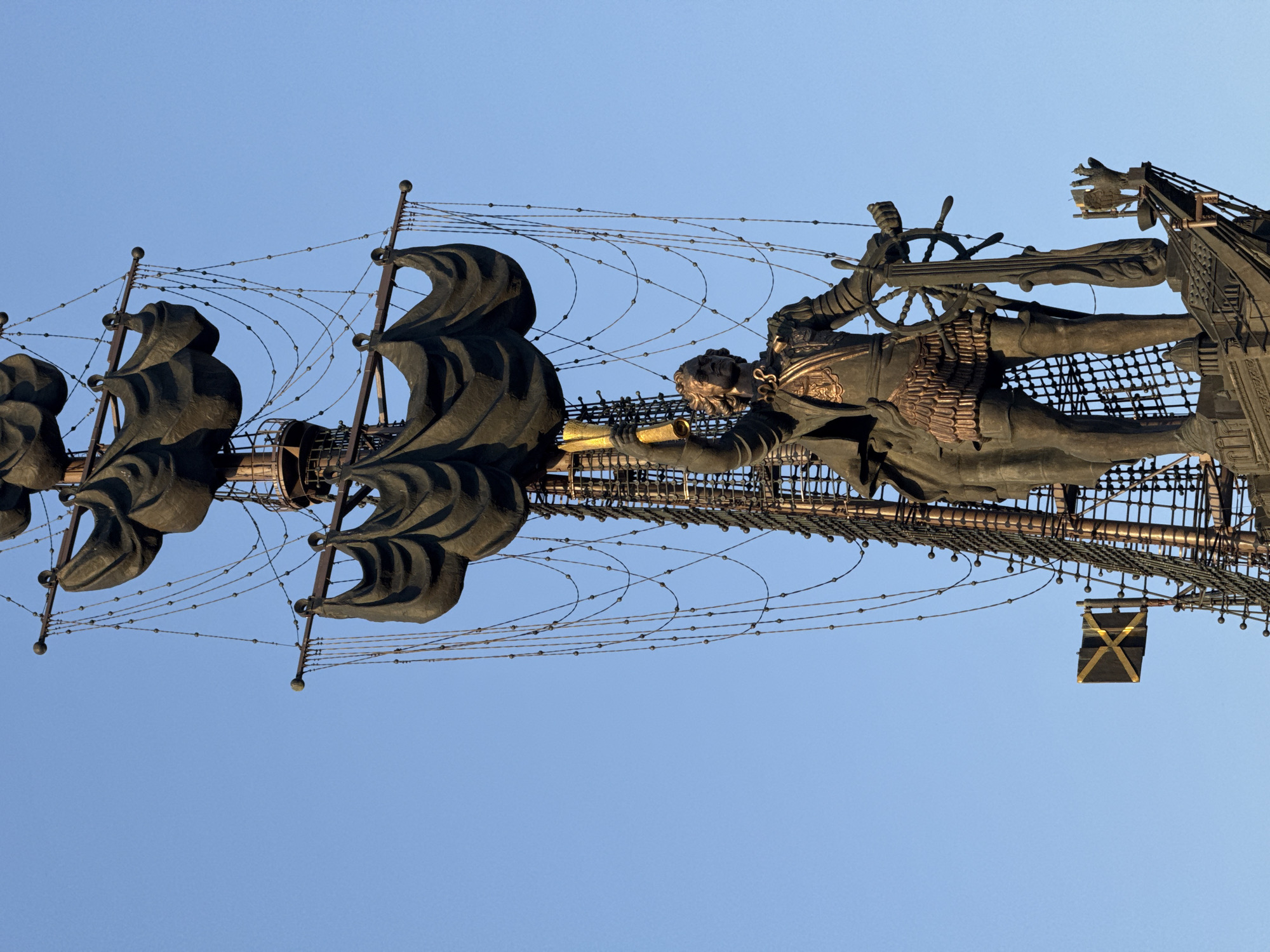 The image shows a monument of Peter the Great, also known as the Peter I monument, located in Moscow, Russia. It depicts a large bronze statue of Peter the Great standing on a ship. He is holding a golden scroll. The ship has several masts with dark billowing sails. The monument is set against a clear blue sky.