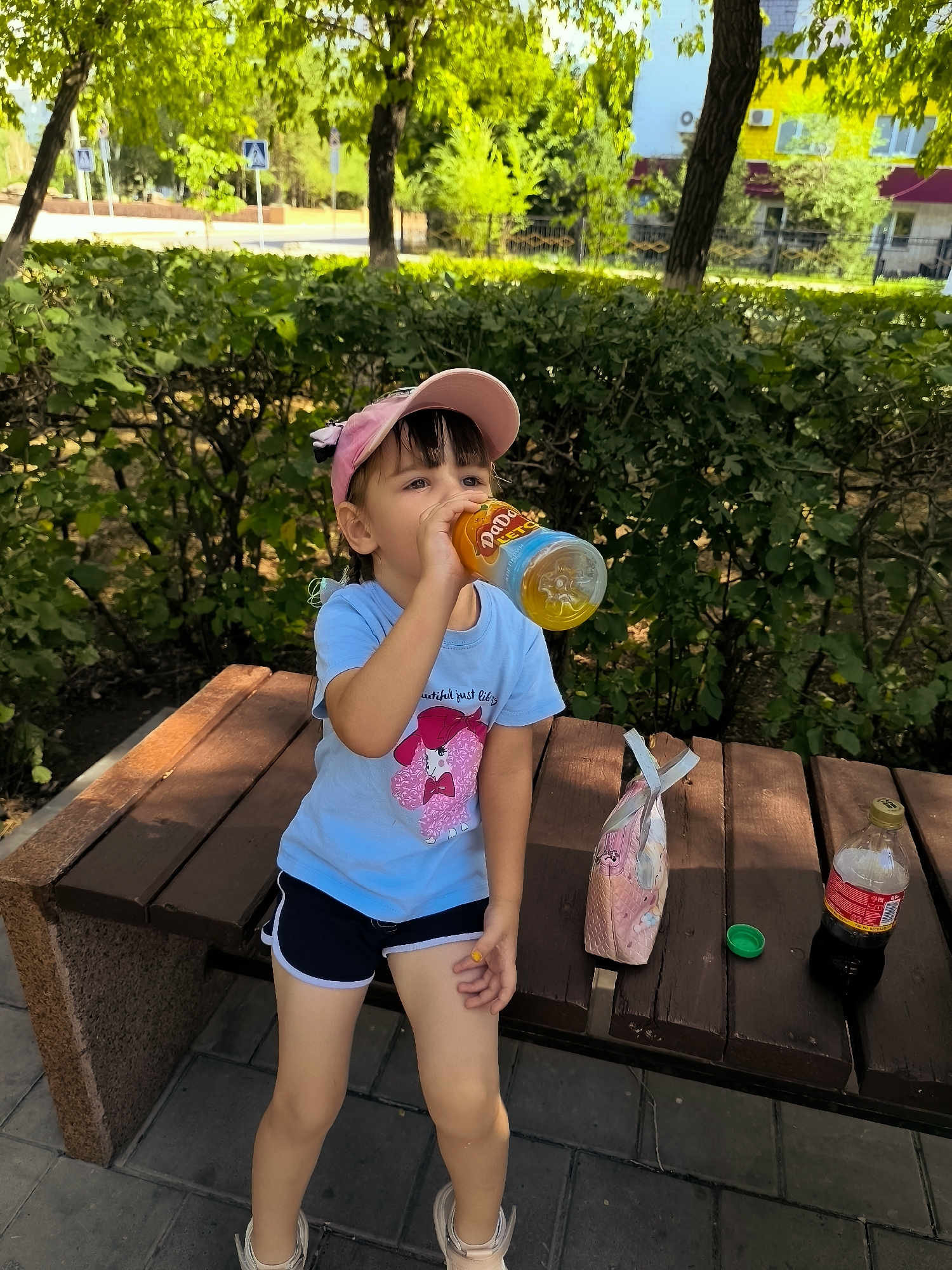In Karaganda, Kazakhstan, a young girl wearing a pink baseball cap and a light blue t-shirt with a graphic print is sitting on a wooden park bench. She is drinking from a clear plastic bottle filled with an orange beverage. Next to her on the bench are a pink patterned bag and a bottle of dark liquid, likely soda, with a green cap lying nearby. The bench is situated outdoors, with lush green bushes and trees forming the background. In the distance, buildings and street signs are visible, suggesting an urban park setting. The girl's shorts are navy blue with white trim, and she is wearing white sneakers. The overall scene depicts a casual moment of refreshment in a park.