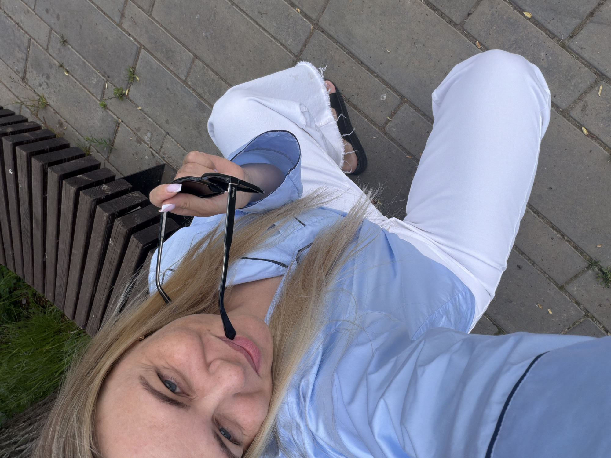 A high-angle selfie captures a blonde woman sitting outdoors in Votkinsk, Russia, beside a wooden slatted bench. She is looking towards the camera with a slight smile, holding a pair of black sunglasses in her right hand. The brand name "Polaroid" is visible on the temple of the sunglasses. She is dressed in a light blue long-sleeved shirt with dark piping along the edges and white wide-leg trousers. Black sandals are visible on her feet. The ground consists of grey rectangular paving stones with small patches of green weeds growing in the crevices. The lighting suggests it is daytime, possibly a mild or overcast day. The scene depicts a casual moment in an urban park or pedestrian area.