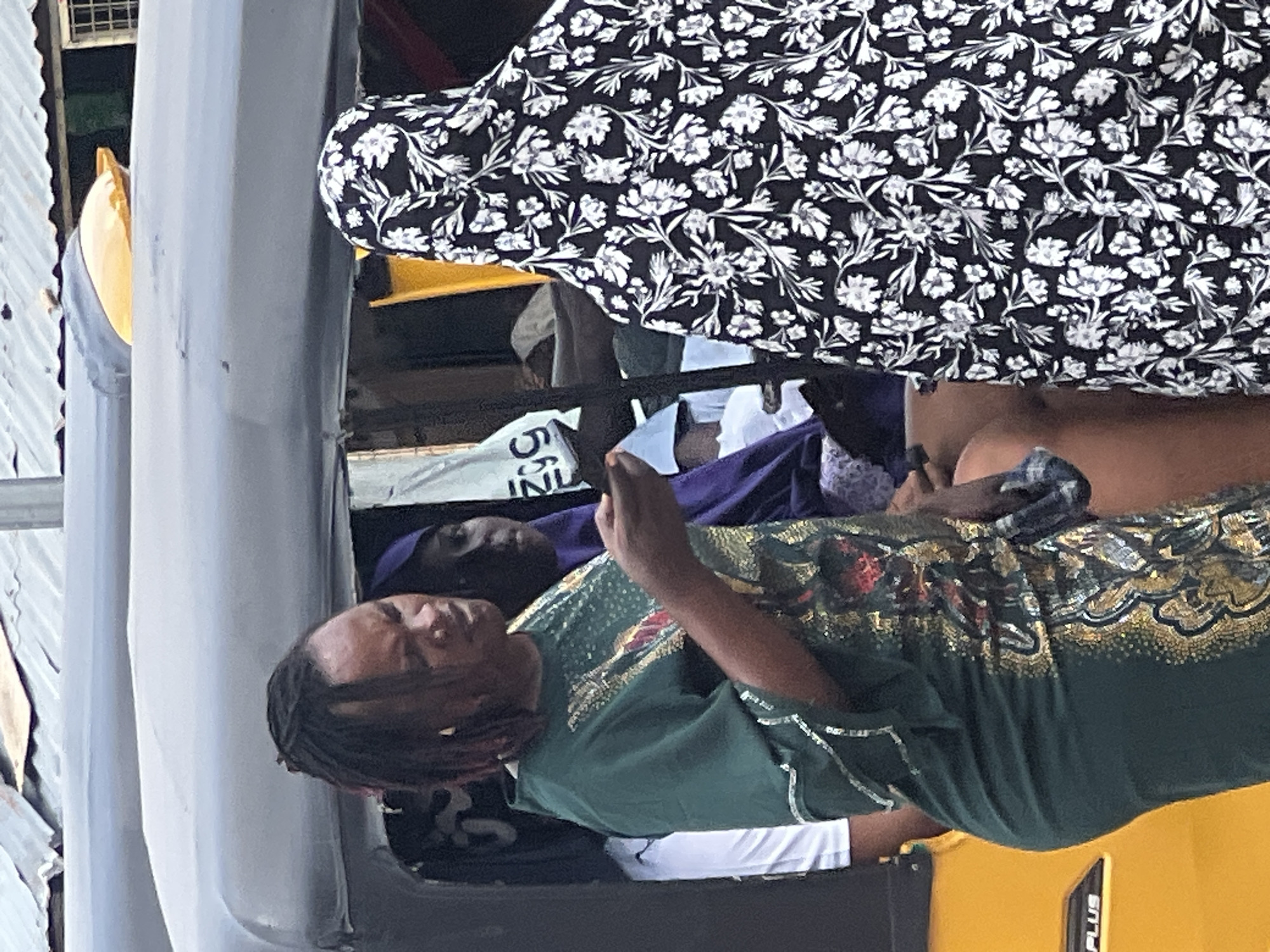 In Jalingo, Nigeria, a group of people stand near a yellow auto-rickshaw. A woman in a green dress with intricate gold and red embellishments on the front is holding a mobile phone. Her hair is styled in dark dreadlocks. Beside her, another person is seen from behind wearing a black and white floral patterned headscarf and covering. A third person, wearing a purple headscarf, is partially visible behind the woman in green. The auto-rickshaw has a dark grey canopy and a yellow front. The background suggests an outdoor urban setting with hints of corrugated metal roofing and some structures.