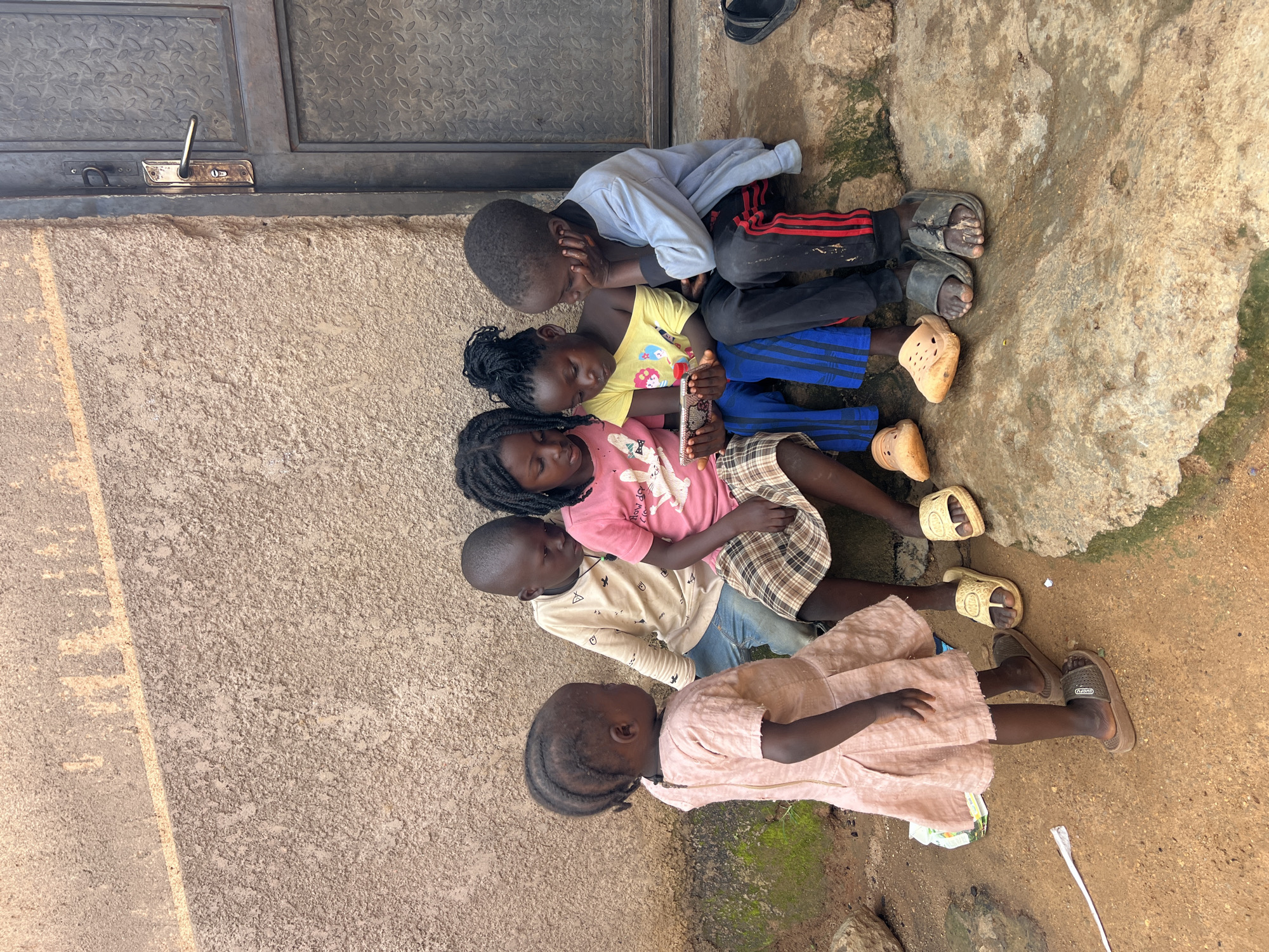 Five children are gathered around a smartphone, seated on concrete steps outside a building with a rough, textured wall and a metal door. The youngest child, a girl in a pink dress, stands to the left, facing away from the viewer. The other four children, three girls and one boy, are seated together, looking at the phone. One girl in a pink t-shirt and plaid skirt is in the center, with two other children on either side of her. A boy in a light blue long-sleeved shirt and black pants with red stripes sits on the far right, resting his head on his hand. The ground is dirt and appears damp in places. The scene is likely in an urban or semi-urban area, potentially within a residential setting. The overall impression is one of shared engagement with technology.