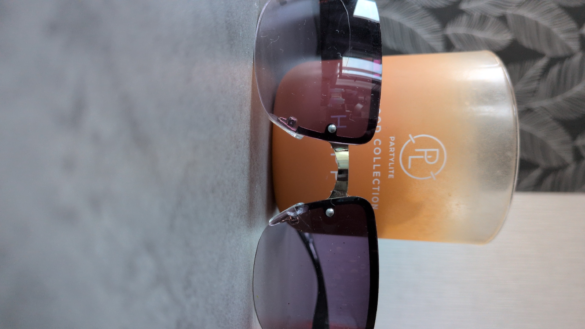 This is a close-up, vertical, eye-level shot of a pair of sunglasses resting on a light-colored, textured surface, possibly a table or countertop. The sunglasses are dark-rimmed with purple lenses. They are positioned diagonally across the frame, with the top lens to the left and the bottom lens to the right.
Behind the sunglasses is a partially visible candle. The candle has an orange gradient color, fading to a lighter shade at the top. The glass jar of the candle is somewhat smudged and dusty. On the side of the candle, there is text printed vertically. The visible text includes "COLLECTION," "PARTY LITE," and a logo featuring the letters "PL" within a circular outline. The background behind the candle is a dark, patterned wallpaper with a light-colored, leafy design.
The lighting appears to be soft, with some reflections visible on the sunglasses lenses, suggesting an indoor setting. There are no people visible in the image. The overall mood is still and neutral. The scene is likely set indoors, given the presence of the candle and the indoor background. The image is cropped tightly on the objects, with the surrounding environment only hinted at by the background pattern. The time of day and weather are not discernible. There are no explicit emotional cues present.