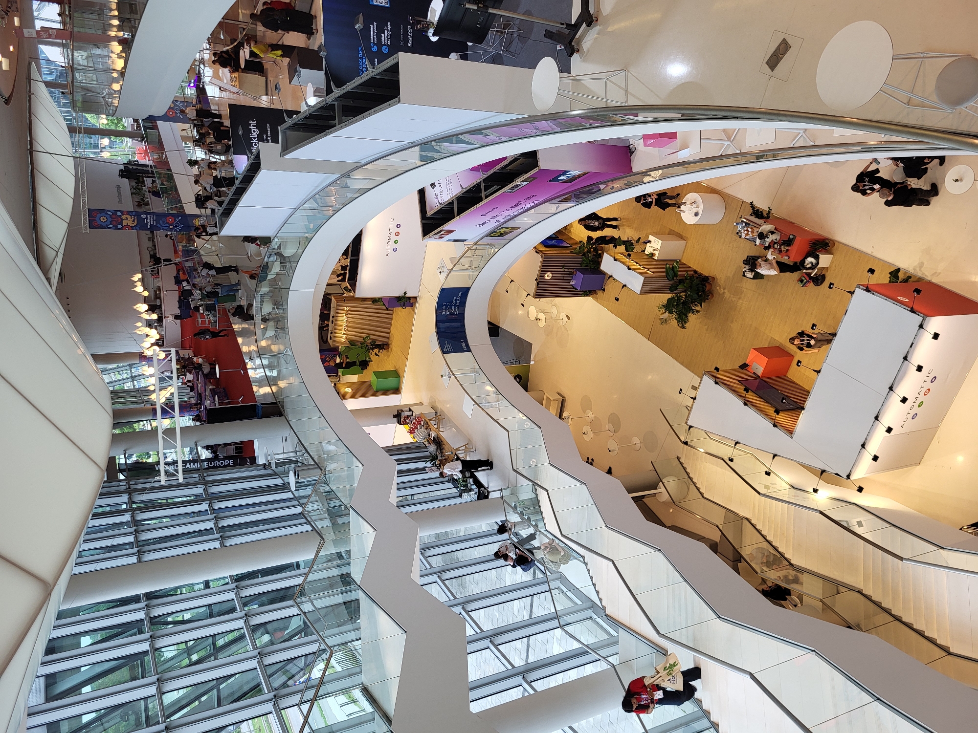 The image displays the multi-level interior of a modern convention center or exhibition hall, located in the city of Unknown, Unknown. The architecture features prominent white curved structures, expansive glass curtain walls on the left revealing an outdoor view of greenery and a road, and a dramatic zig-zagging staircase with glass balustrades connecting several floors.
The building is actively hosting an event or conference. Numerous temporary booths and display areas are set up across the different levels. On the lowest visible floor, a prominent white and orange/red booth for "AUTOMATTIC" is visible, with colorful circular logos beneath its name. Further up on a curved balcony, a sign for "ticklight." is partially seen. Other signage indicates various zones like "Track 1," "Explo. Zone," and "Confer. Zone," along with a banner for "RESTAURACJA" (restaurant in Polish) decorated with folk art patterns on an upper level. Another partially visible sign reads "TEAM EUROPE."
Dozens of individuals populate the space. People are seen walking up and down the stairs, congregating around booths, standing at tall white tables, and interacting with staff. One person in a red top and dark pants, carrying a canvas bag, is descending the stairs in the foreground. Several staff members are visible, including baristas at a coffee station on a lower level and personnel behind a buffet-style food and drink setup on an upper floor. The atmosphere appears busy with attendees mingling and engaging with the exhibition content.
The interior is brightly lit by a combination of natural light filtering through the large windows and artificial lighting, including decorative pendant lights hanging from the high, fabric-paneled ceiling. Potted plants are strategically placed near some booths for decoration. The overall impression is one of a spacious, well-maintained venue bustling with activity.