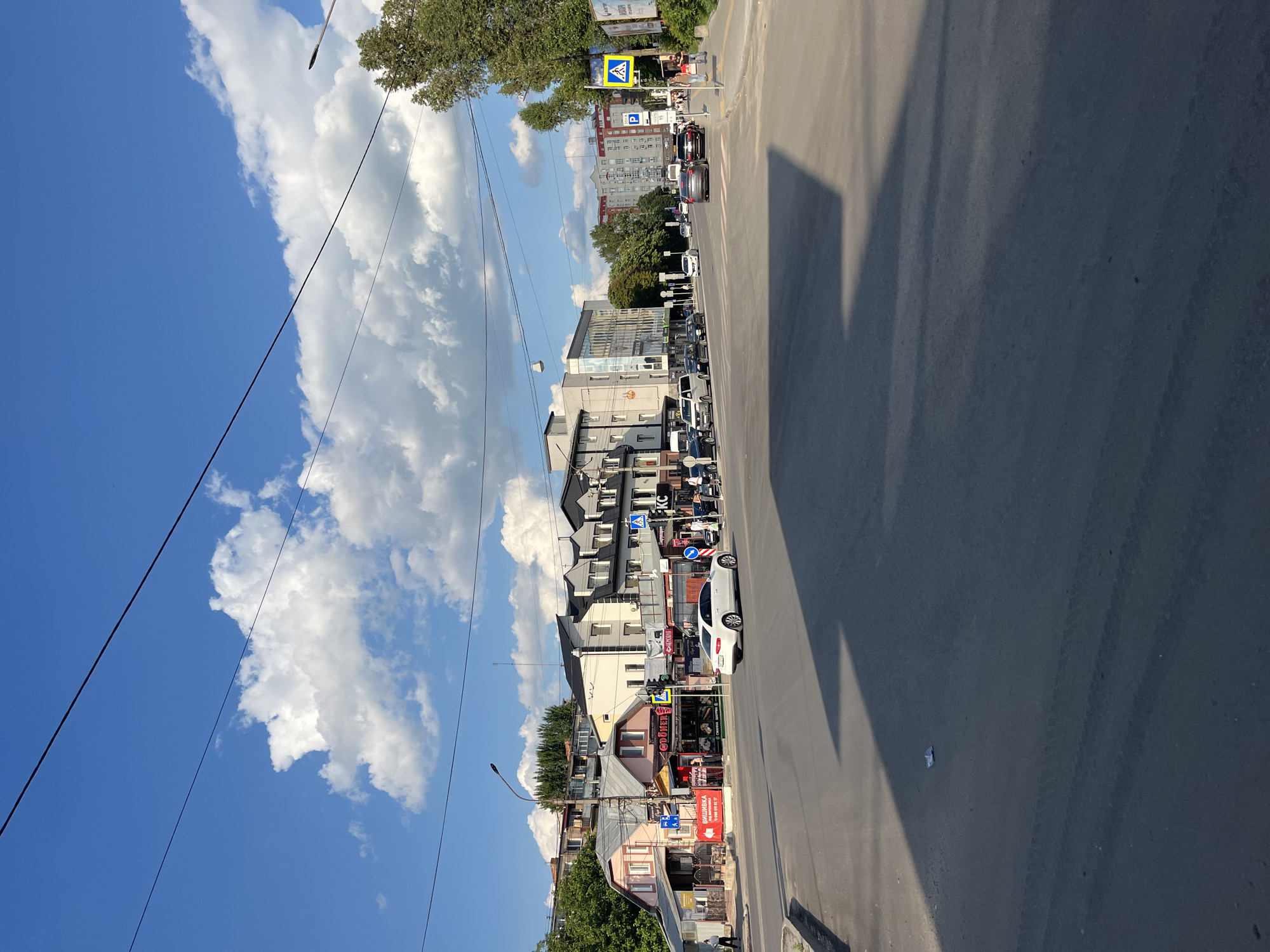 This is a daytime, eye-level, medium-shot photograph of a street intersection in Ternopil, Ukraine. The foreground shows an asphalt road with tire tracks and a prominent shadow cast from the left. In the middle ground, a white sedan is turning left, and other cars are present in the lanes. Pedestrians are visible on the sidewalks. The background features a row of buildings of various architectural styles and heights, including a two-story pink building with commercial establishments on the ground floor and residential units above, and a modern, multi-story building with glass facades. A street light is visible to the left of the pink building. A crosswalk with a pedestrian signal is located to the right of the pink building. Further in the background, more residential buildings and trees line the street. The sky is blue with scattered white clouds.