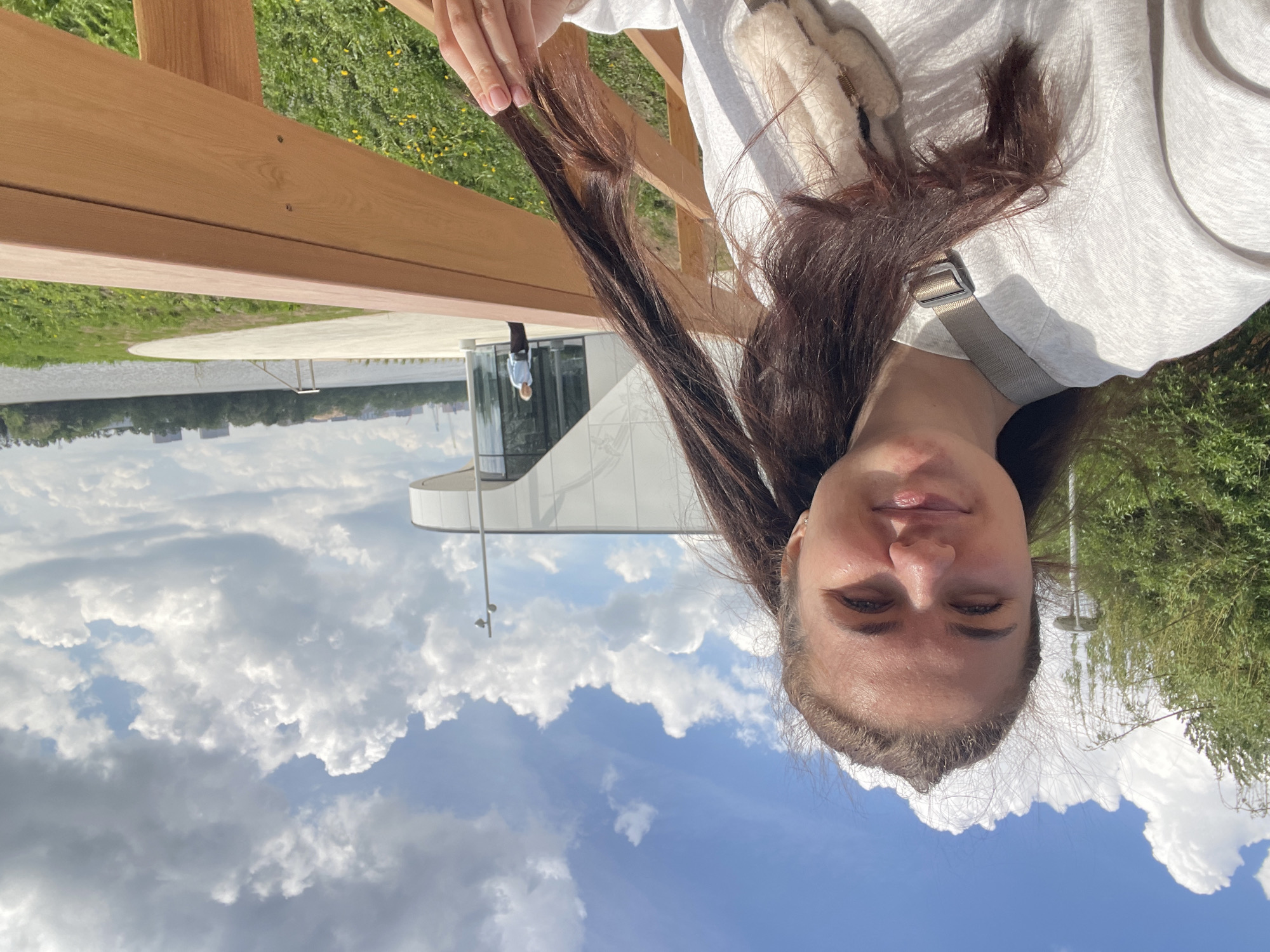 A woman with long brown hair is holding a strand of her hair near a wooden railing outdoors. She is wearing a light-colored top and a bag strap is visible on her shoulder. Behind her, there's a modern building with large glass windows and white curved walls. In the background, a body of water is visible with trees and buildings on the opposite shore. Above, there's a blue sky with white clouds. The scene is located in the city of Moscow, Russia.