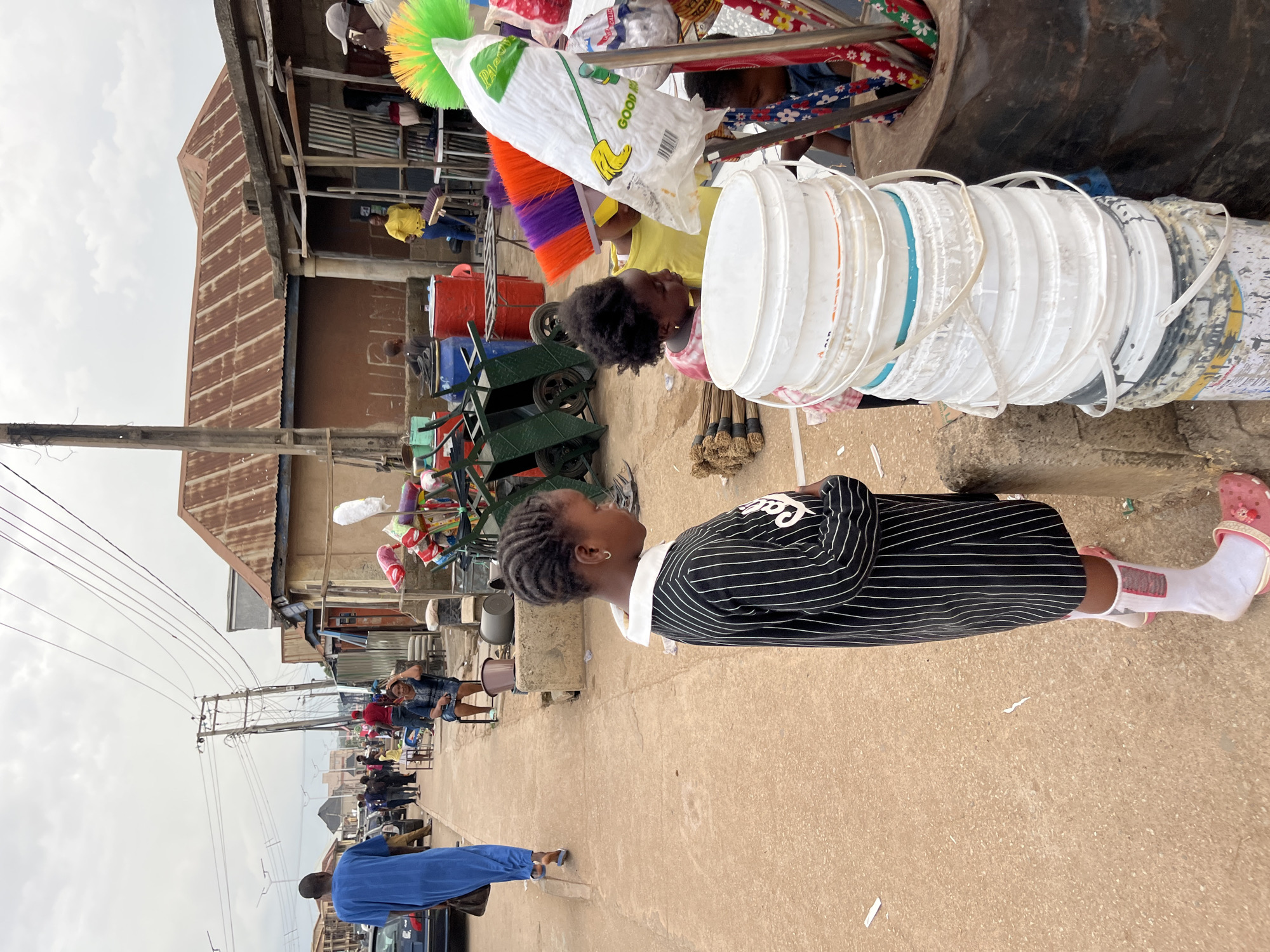 The image depicts an outdoor marketplace scene in Bukuru, Nigeria, under an overcast sky. A young girl, wearing a black and white striped dress, white socks, and pink clogs, stands in the foreground, facing a stack of white buckets. Her hair is styled in braids. Behind her, a younger child in a yellow top looks at a mop. Several brooms with colorful handles are visible on the ground. To the left, a man in a blue robe walks away from the camera, carrying a brown bag. Other people and stalls line the dusty street. Structures with corrugated metal roofs and rough wooden supports are present in the background. The scene is typical of a bustling Nigerian urban market.