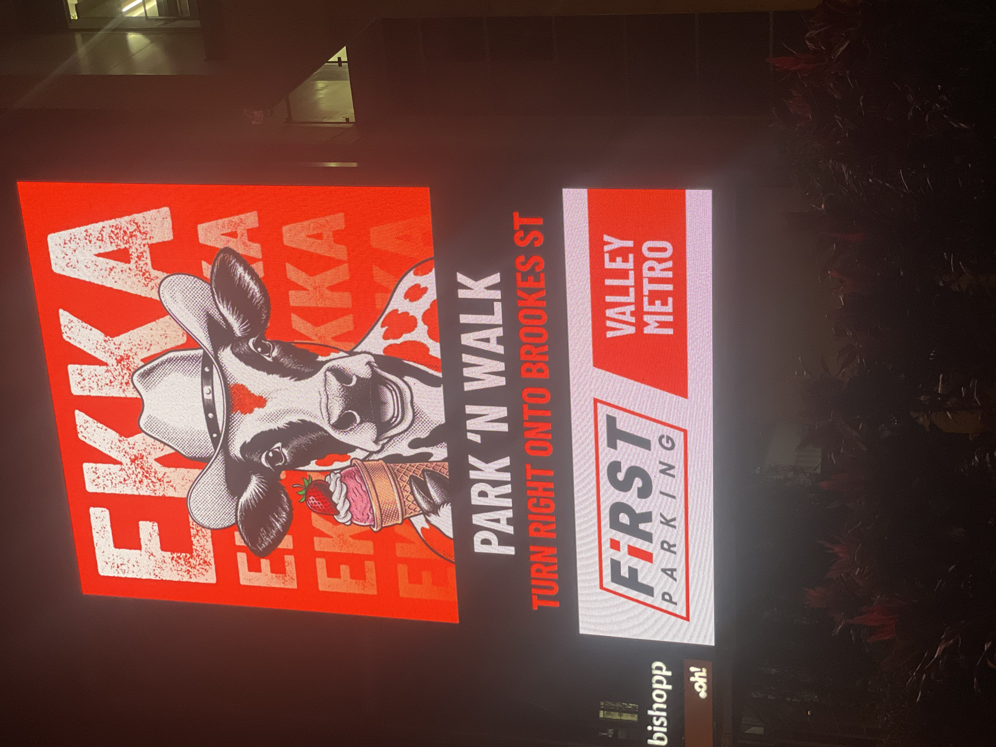 This is a nighttime photograph of a large digital billboard in Brisbane, Australia, likely located in the vicinity of the Ekka grounds and the Valley Metro precinct, judging by the signage. The billboard displays vibrant, illuminated content against a dark background, suggesting it is a prominent advertisement.
The top section of the billboard features a large, stylized graphic of a cow wearing a cowboy hat and holding an ice cream cone with a strawberry on top. Behind the cow, the word "EKKA" is repeated multiple times in large, red, distressed lettering.
Below this graphic, white text reads "PARK 'N WALK," and red text below that says "TURN RIGHT ONTO BROOKES ST." This indicates directions for parking or traffic.
The bottom section of the billboard is divided into two panels. The left panel displays the logo for "FIRST PARKING," featuring the word "FIRST" in a stylized red font with a gray outline and "PARKING" in gray text below. To the left of this logo, a partial view of a brown sign with white text "bishopp" and a small orange logo with "oh!" is visible. The right panel contains the words "VALLEY METRO" in large white letters against a red background.
The overall scene is dark, with the illuminated billboard being the primary light source. Below the billboard, the faint outlines of foliage and possibly some decorative lighting can be seen, suggesting an outdoor public space. The image quality is slightly blurred, indicating motion or low light conditions.