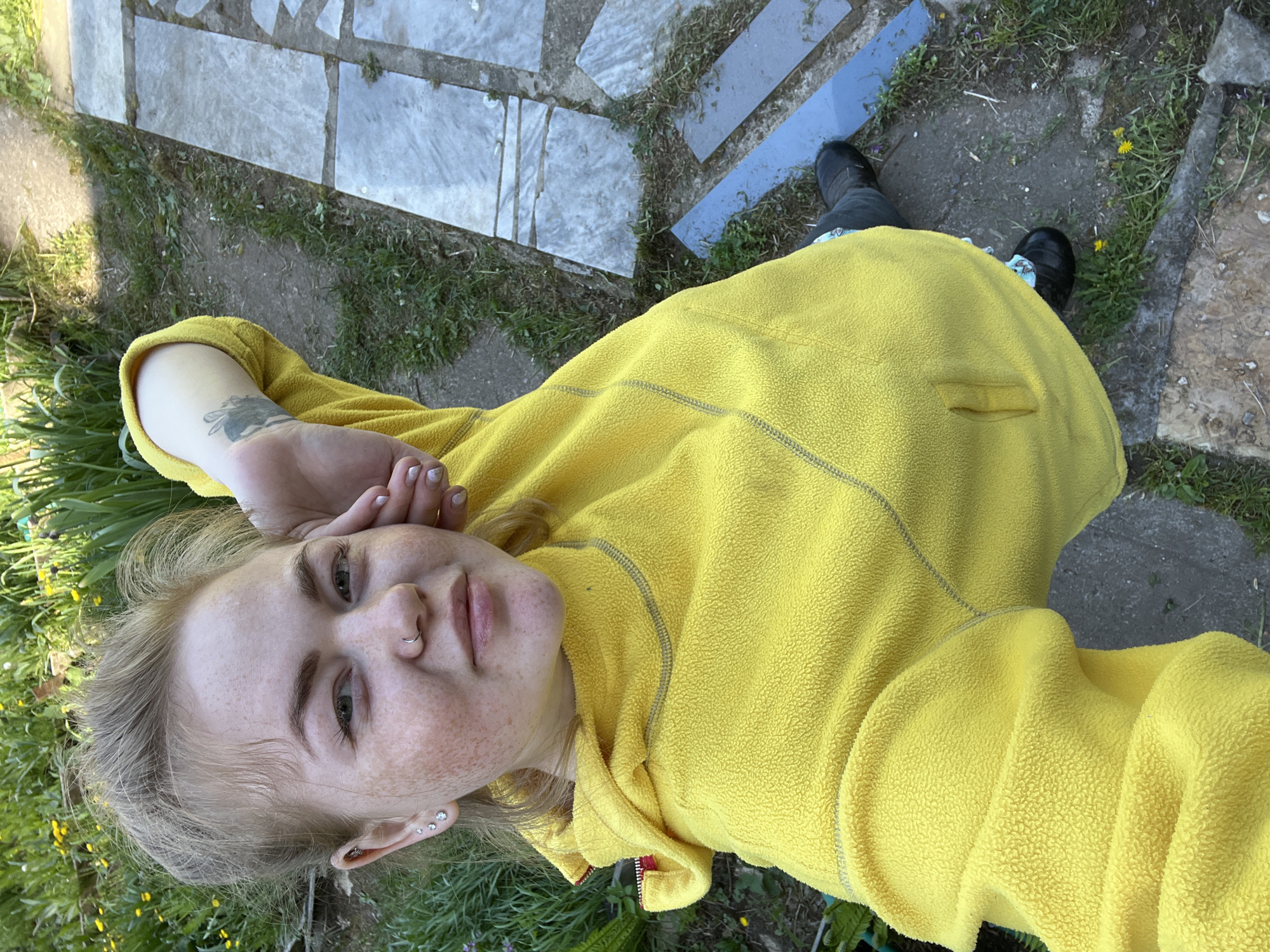 A young woman with fair skin, freckles, and blonde hair is lying on the ground, possibly on grass, near a stone or concrete pathway in Chaikovsky, Russia. She is wearing a bright yellow fleece jacket and dark shoes. Her left hand is resting near her face. The environment consists of grass, weeds, and paving stones or concrete slabs. A piece of blue material is visible near her feet.