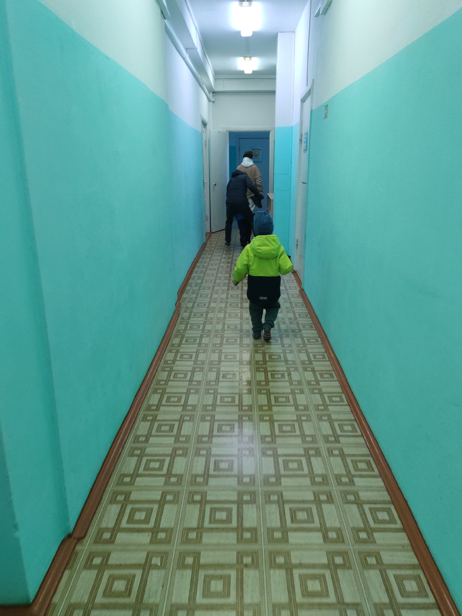 The image depicts a long, brightly lit indoor hallway, likely within a public or residential building in Chaikovsky, Russia. The walls are painted in two distinct horizontal sections: a vibrant turquoise or teal color on the lower half, and a lighter off-white or cream color on the upper half, meeting at a clean line. A dark brown baseboard runs along the bottom of the walls. The floor is covered with linoleum or tiles featuring a repetitive geometric pattern of interconnected squares in shades of beige and brown. Several simple ceiling light fixtures illuminate the corridor.
In the foreground, a young child, viewed from behind, walks down the hallway away from the camera. The child wears a bright lime green hooded jacket, dark blue or black trousers, and a dark blue beanie. Further down the hallway, two other individuals, also seen from behind, are walking in the same direction. One appears to be wearing a light-colored (possibly beige) hoodie or jacket, while the other wears a darker jacket. The overall scene suggests a routine passage through a utilitarian indoor environment. No legible text is visible within the frame.