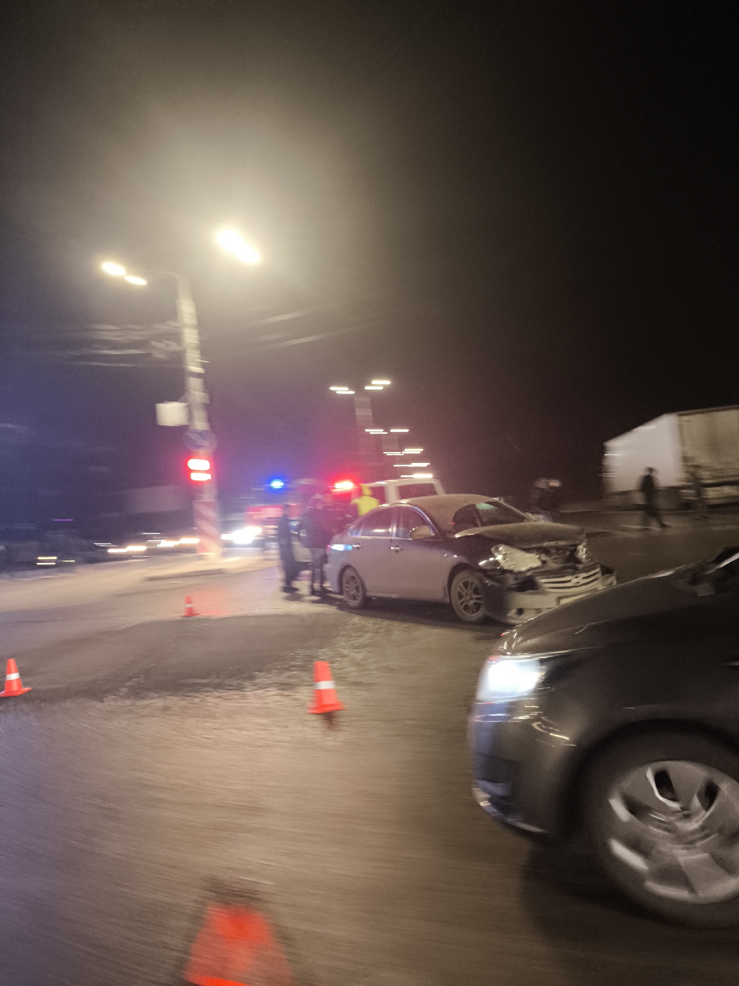 The image depicts a car accident scene at night in what appears to be an urban area. Several orange traffic cones are placed around the site. Two cars are visibly damaged; one is a silver sedan with significant front-end damage, and the other is a dark-colored car with a visible headlight. Several people stand near the silver car, possibly involved in the accident or assisting. Emergency lights from a vehicle, likely a police car or ambulance, are flashing in the background. A red traffic light is also visible. In the background, there is a large white truck. Based on the general appearance and urban setting, the image shows a car accident in an unidentified city. The information about the city of Izhevsk, Russia, cannot be verified from the provided image.