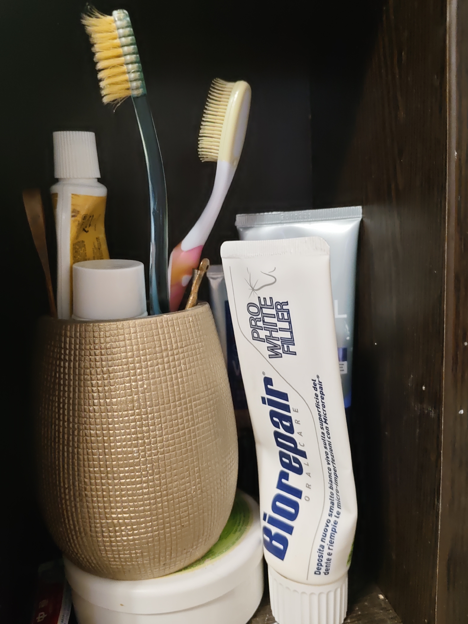 The image shows a close-up of a bathroom shelf or cabinet. The foreground is dominated by a textured, golden-colored holder containing several toothbrushes and tubes of toothpaste. Two toothbrushes are visible, one with green and white bristles and a blue handle, and another with yellow bristles and a white handle. There are also tubes of toothpaste, one of which is a Biorepair toothpaste tube with "PRO WHITE FILLER" written on it in large blue letters. Behind this, another tube of toothpaste, appearing to be silver, is partially visible. The background consists of a dark, wooden surface, likely the interior of a cabinet or shelf. The lighting suggests an indoor environment, possibly a bathroom. There are no people or animals visible in the image, and no specific geographical markers are present to indicate a location in Rostov-on-Don, Russia, or any other specific city.