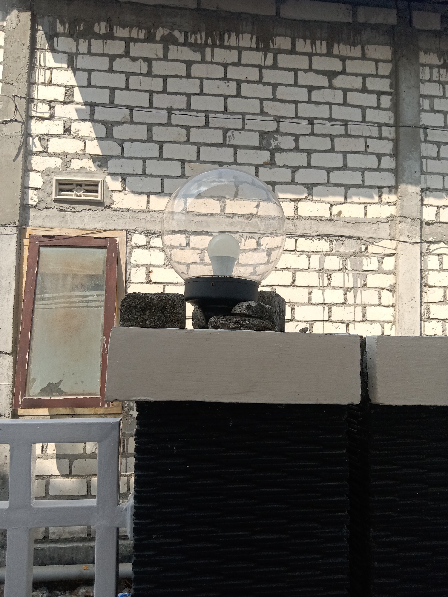 A clear, medium-shot, vertical photograph taken outdoors on a sunny day shows a decorative outdoor light fixture mounted on a wall in Klepek, Indonesia. The fixture consists of a transparent, spherical glass globe with a white LED bulb inside, resting on a black base. The base is supported by several dark, rough-textured stones, which are positioned on top of a white ledge. This ledge extends across the top of a black, textured wall. To the left of the light fixture, a small, weathered window with a brown frame is set into a white brick wall. The white brick wall has a rough concrete finish at the top, with visible dripping cement. A thin wire runs horizontally across the brick wall. In the foreground, a section of a light grey fence with square openings is visible. The background is dominated by the white brick wall, which shows signs of weathering and uneven construction. Sunlight casts distinct shadows on the wall, highlighting the texture of the bricks and the mortar.