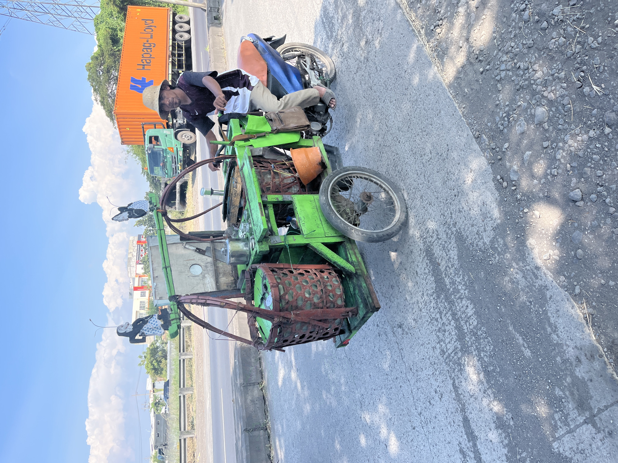 In Pamotan, Indonesia, a man wearing a straw hat and a dark t-shirt rides a three-wheeled motorcycle with a green wooden cart attached. The cart is equipped with a large bamboo basket filled with green items, a metal container, and an orange bucket. Two decorative figures, one resembling a puppet and the other a butterfly, are attached to the front of the cart. In the background, a large orange Hapag-Lloyd shipping container truck drives on a highway, with lush green trees and buildings visible on the side. The sky is clear blue with fluffy white clouds. The motorcycle and cart are on a paved road with patches of gravel and debris.