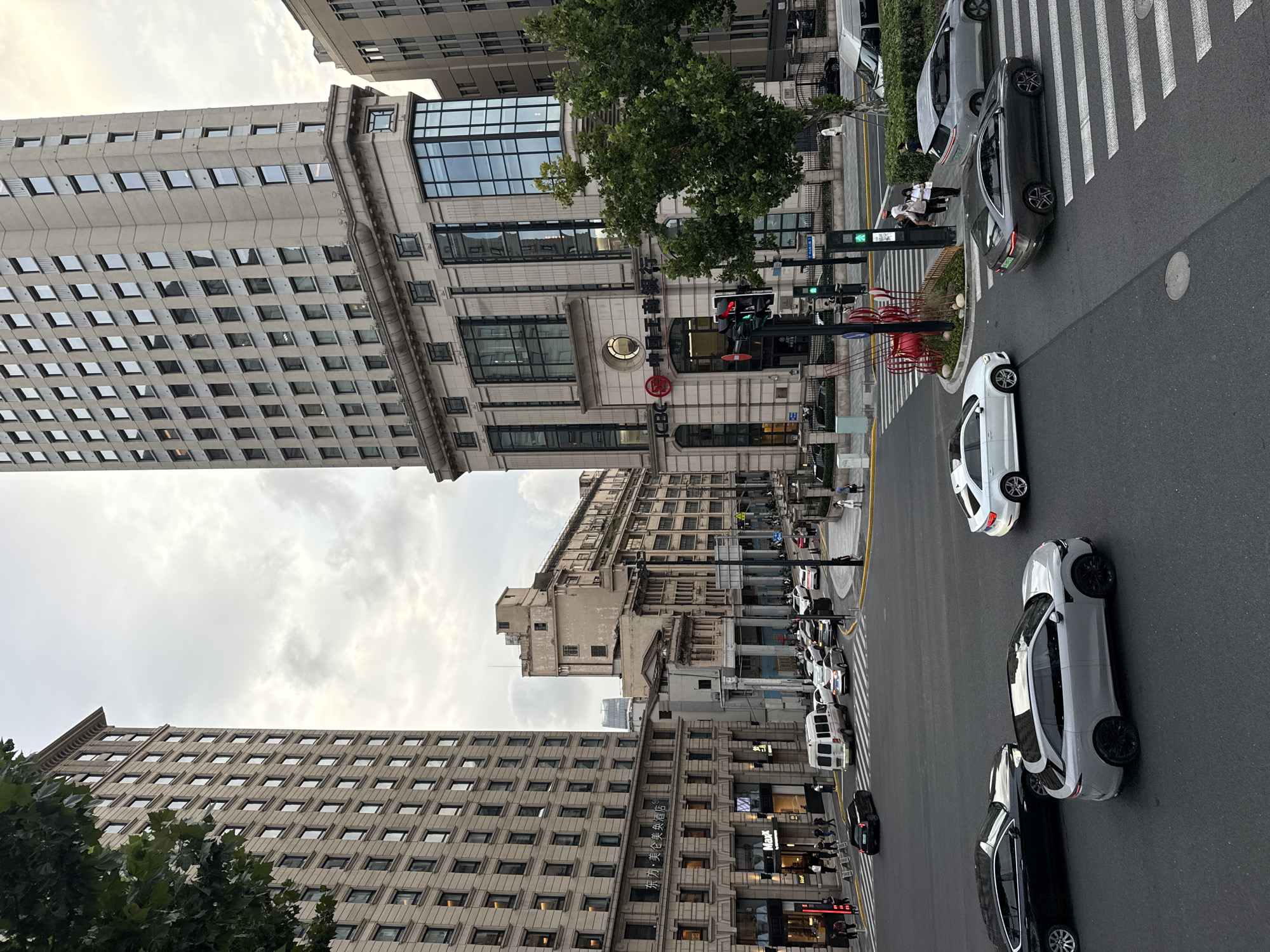 The image captures a street intersection in Shanghai, China, likely in an older district given the architectural styles of the buildings. Two prominent buildings dominate the scene: a tall, modern high-rise on the right with a distinctive grid of windows and a facade featuring blue-tinted glass sections, and a slightly shorter, older building on the left with a more classical facade and numerous uniformly spaced windows.
The intersection itself is busy with vehicular traffic. Several cars are visible, including a light gray SUV in the foreground, a black sedan to its left, and a dark gray sedan on a crosswalk to the right. White passenger cars and a white van are also present in the middle ground, navigating the intersection. Pedestrians are seen on the sidewalks, with a few individuals walking or standing near crosswalks.
Traffic lights are clearly visible and appear to be functioning, with some indicating green. The street is paved with asphalt and marked with white lane dividers and crosswalks. In front of the modern building on the right, there is a decorative red sculpture. Green trees are scattered throughout the scene, adding a touch of nature to the urban environment. The sky is overcast, suggesting a cloudy or hazy day. Signs with Chinese characters are visible on some buildings, confirming the location in China. The overall impression is one of a bustling urban environment in Shanghai.