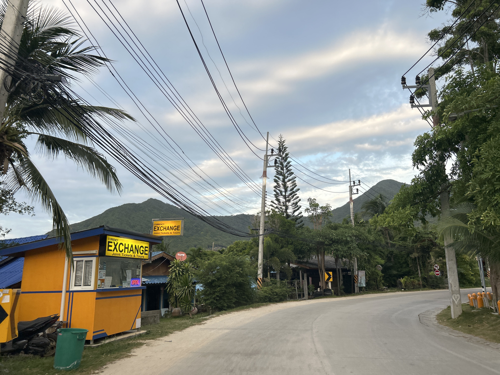 The image shows a street scene in the Ko Pha-ngan District of Thailand, likely in a semi-tropical area given the vegetation. A bright yellow currency exchange booth with a blue roof is prominent on the left side of the road, displaying a yellow sign that reads "CURRENCY EXCHANGE" with "Joint Tickets & Tours" underneath. A motorcycle is parked next to the booth, and a green trash bin is situated in front of it.
The road curves gently to the right and appears to be paved with a light-colored material, possibly gravel or a similar surface. On the right side of the road, there are more trees and what appear to be small buildings or stalls. Overhead, a dense network of power lines stretches across the sky, supported by concrete utility poles.
In the background, lush green mountains rise up, partially obscured by trees and foliage. The sky is overcast with scattered clouds, suggesting either early morning or late afternoon. The overall impression is a somewhat remote and tropical location, possibly a tourist area given the presence of the currency exchange booth. There are no people visible in the image.