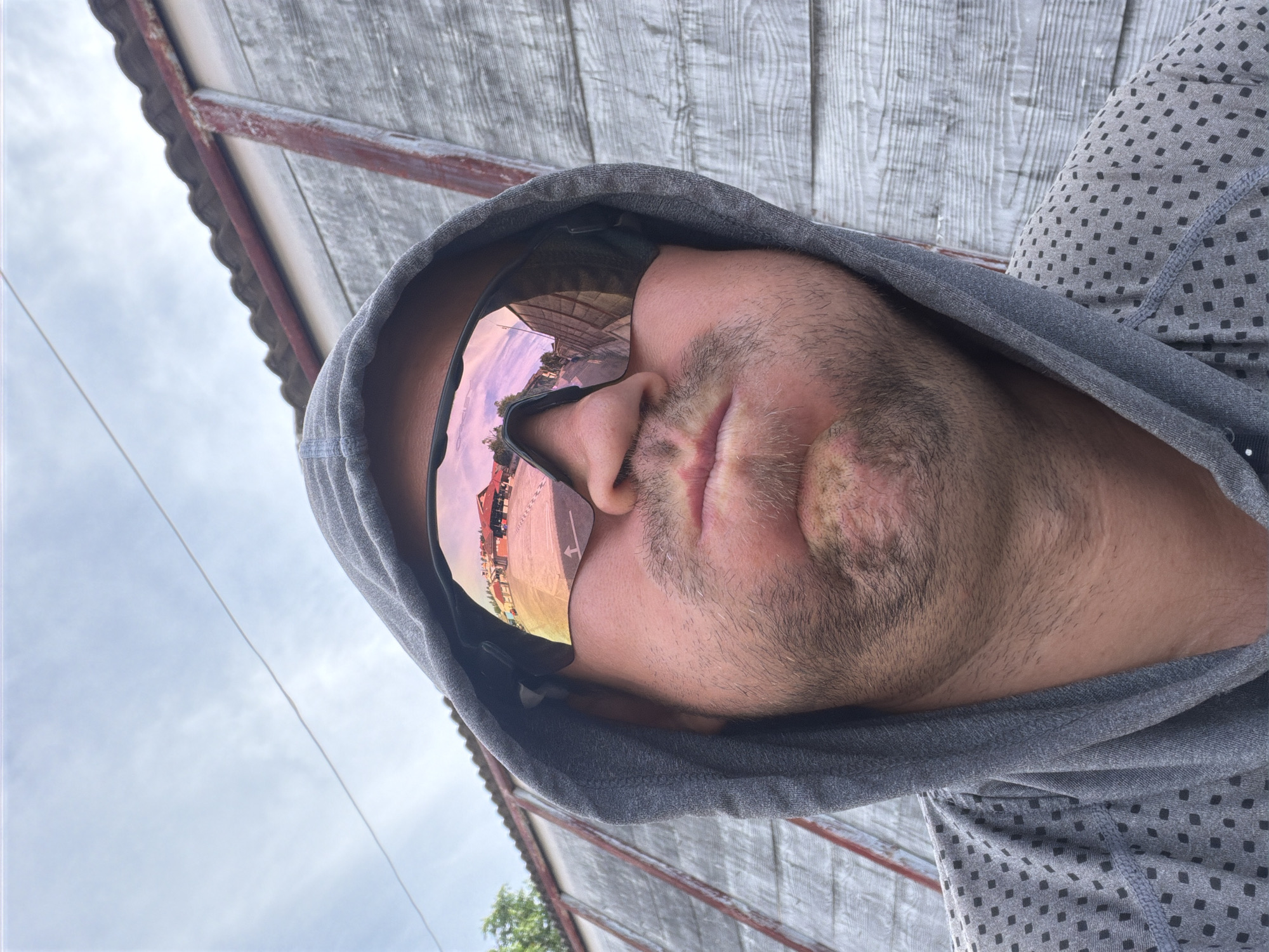 A low-angle, close-up, selfie-style image shows a person wearing a grey hooded sweatshirt and mirrored sports sunglasses, obscuring their eyes. The person's face is puffed out, with their lips pursed. The sunglasses reflect a scene of buildings with red roofs and a street, indicative of a market or town center. The background consists of a textured, weathered wooden wall with red trim and a cloudy sky. The overall setting suggests an outdoor environment in Baan Mae Nam, Thailand.