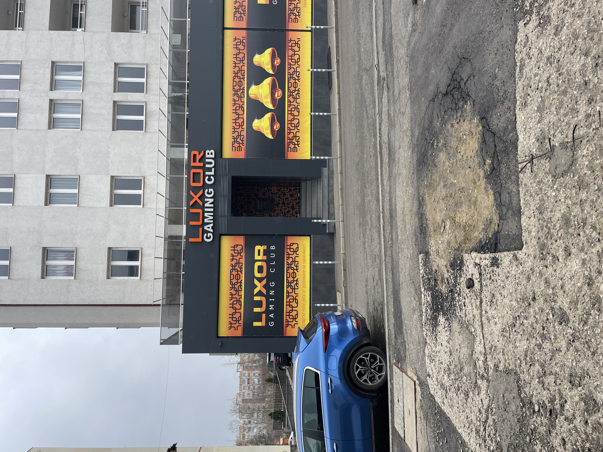 The image captures an exterior view of the "Luxor Gaming Club," located in Sofia, Bulgaria. The club's facade is prominently displayed with its name in orange letters above the entrance. The building is multi-storied, with a gray facade. A blue car is parked on the side of the street in front of the club. The street and sidewalk appear worn and cracked. The sky is overcast.