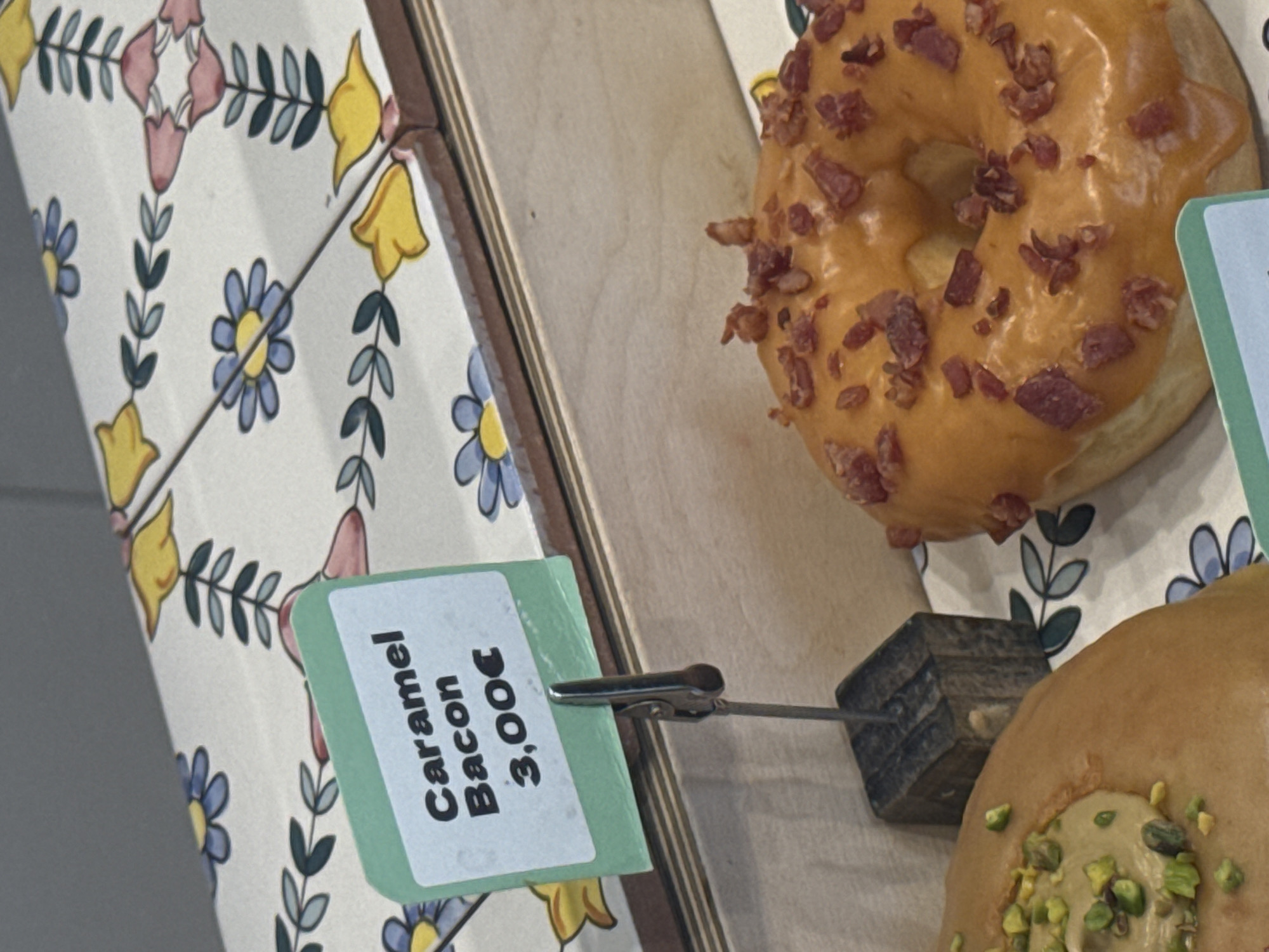This image captures a close-up, high-angle shot of a display of gourmet donuts, likely in a bakery or cafe in Milan, Italy, given the Euro currency. The scene is brightly lit, showcasing the textures and colors of the pastries.
The primary subject is a donut with an orange, glossy glaze generously sprinkled with numerous small, reddish-brown pieces of crispy bacon. This donut is positioned in the upper right quadrant of the frame. Below it, partially visible in the bottom right corner, is another donut featuring a light brown glaze, topped with chopped green pistachios.
A light green and white price tag, attached to a small wooden block by a metal clip, is prominently displayed in the bottom left. The text on the tag is rotated counter-clockwise and clearly reads: "Caramel Bacon 3,00€". This tag corresponds to the bacon-topped donut.
The donuts are set against a decorative surface, possibly tiled or fabric, featuring a repeating floral pattern. This pattern includes blue and yellow bell-shaped flowers, pink blossoms, and green leaves on a white background. A wooden border or shelf edge separates this decorative surface from another plain wooden surface where the donuts are displayed. The overall impression is one of tempting desserts showcased in a charming setting.