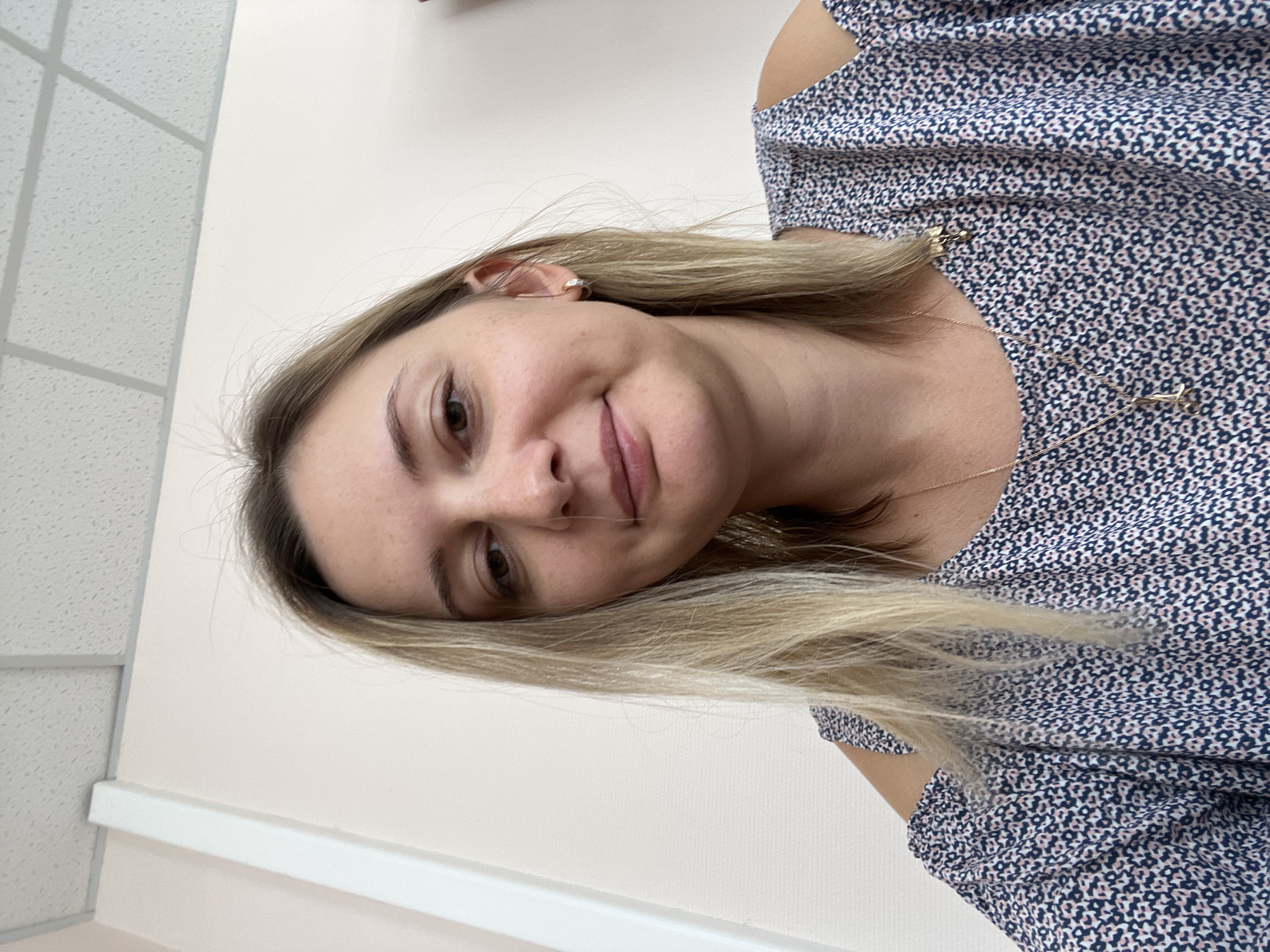 A medium shot photograph shows a fair-skinned woman with shoulder-length blonde hair and a blue and white patterned off-the-shoulder top. She is looking directly at the camera with a slight smile, her right eye slightly closed. The background is a plain, light-colored wall and a white ceiling with acoustic tiles, typical of an indoor office or meeting room. The woman is wearing a small gold hoop earring in her visible right ear and a delicate gold necklace with a small scissors charm. Her makeup is minimal, highlighting her natural features. The lighting appears to be from overhead fluorescent lights, creating a neutral and evenly lit environment. The image seems to be a selfie, taken at eye level. There is no indication of the specific location within Volgograd, Russia, beyond it being an indoor setting.