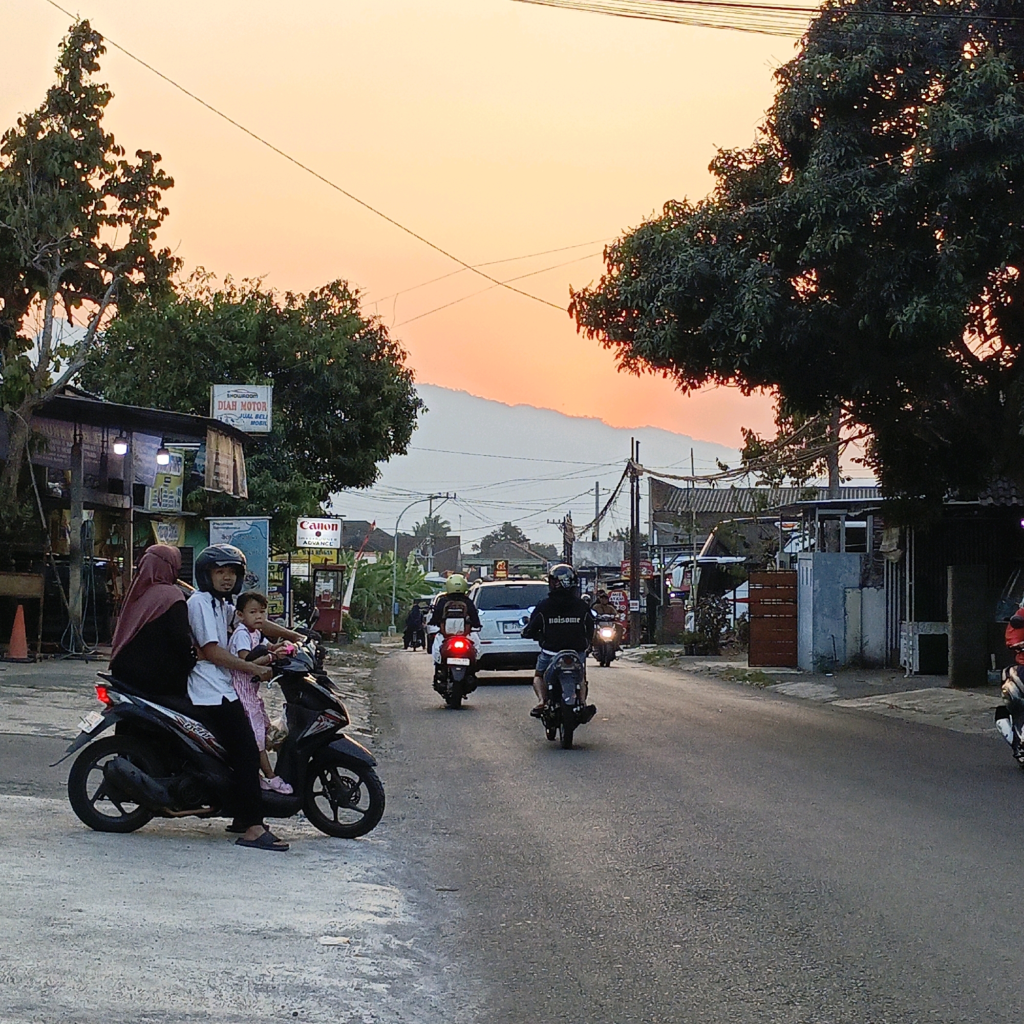The image depicts a street scene in Gedangan, Indonesia, during sunset. The sky displays a gradient of orange and yellow hues. In the foreground, a family of three, a woman wearing a hijab, a man, and a child, are riding on a black motorcycle. The man is driving, wearing a helmet, and the woman is seated behind him. The child is in front of the woman, also holding onto the man. They are on the left side of a paved road.
Along the roadside, there are buildings, including what appears to be a small shop or gas station with signs, one of which reads "DIAH MOTOR JUAL BELI MOBIL" (Diaz Motor Buy Sell Car) and another with a Canon logo. Trees with dense green foliage are visible on either side of the street, with one large tree dominating the right side of the frame, its leaves backlit by the setting sun.
Several other motorcycles are on the road. One motorcycle is directly behind the family's motorcycle, with a rider wearing a black jacket with the word "naitsome" on it. Further down the road, a white car is visible, and more motorcycles are seen in the distance. Utility poles with numerous wires crisscross the sky above the road, suggesting the presence of power and communication lines. The overall atmosphere suggests a typical evening commute or activity in a local Indonesian town.
