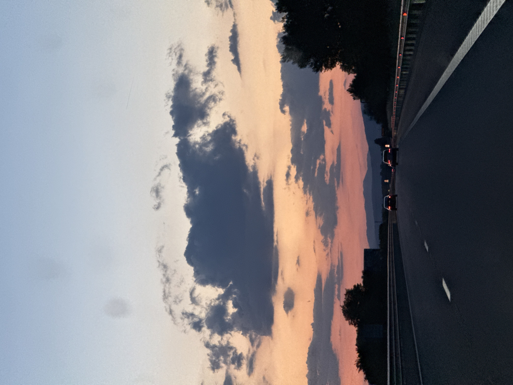 The image depicts a view from a highway at dusk. The sky is filled with clouds, transitioning from a dark gray to a soft orange and pink as the sun sets. A dark, dense cloud formation is prominent on the left. The right side of the image shows a highway with multiple lanes. Guardrails are visible along the edge of the road. Several cars can be seen driving in the distance. Trees line the side of the highway in some areas. The image does not contain enough information to know the location in the city of Saraia, Bulgaria.