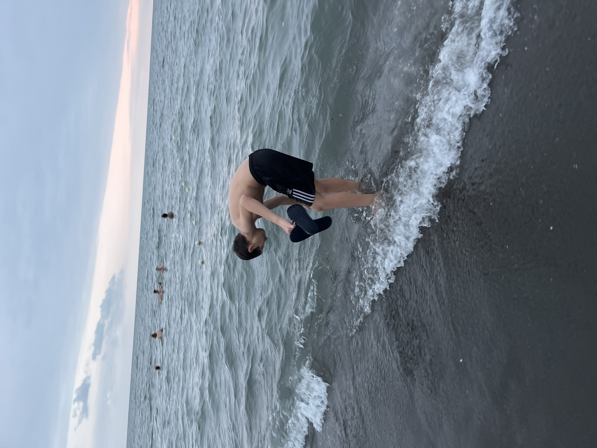 The image shows a young boy in black shorts, backbending near the shoreline of a body of water. He appears to be holding black flip-flops in his hands. The water is a murky gray with white foamy waves crashing near his feet. The sky above is a pale blue with hints of pink and gray clouds, suggesting it may be dawn or dusk. In the distance, there are several other small figures in the water. The scene appears to be a beach or lakefront, given the presence of water and the shoreline. The boy's posture suggests a playful or athletic action, possibly a gymnastic move or simply enjoying the water. There is no visible text in the image. The location is Ureki, Georgia.