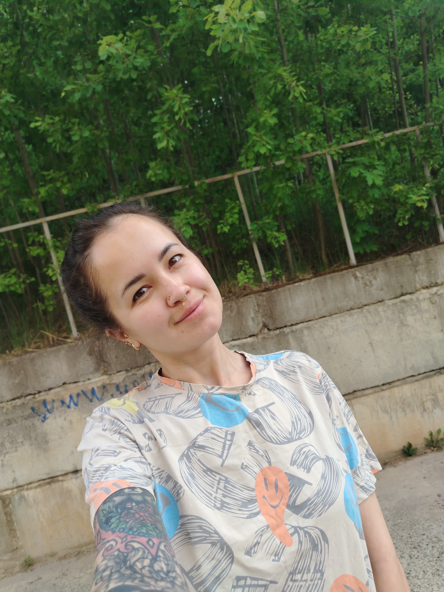 A woman takes a selfie outdoors in a concrete and green space. The woman has dark hair pulled back, a nose ring, and earrings. She wears a printed t-shirt and has a tattoo on her arm. Behind her, a concrete retaining wall runs along the bottom of the frame. Above the wall is a metal railing and a dense thicket of green trees. In the bottom left corner, graffiti is visible on the wall. The location is near Noviy, Russia.