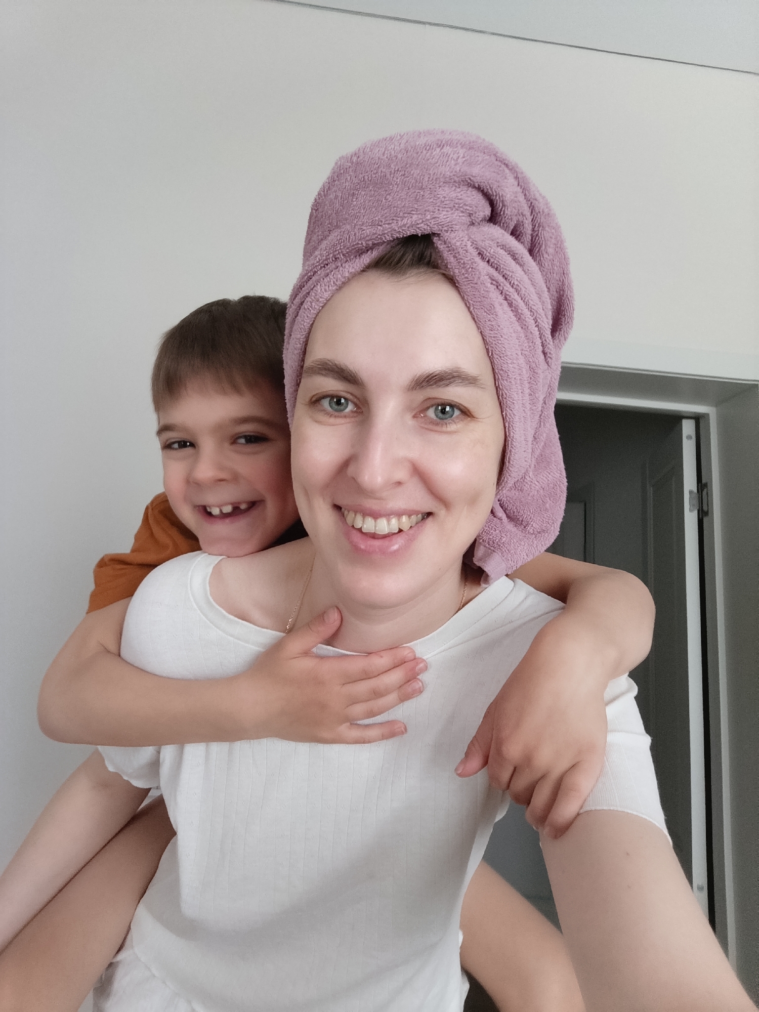 A smiling woman with a lavender towel wrapped around her head is giving a piggyback ride to a smiling boy, who is wearing an orange shirt. She is wearing a white top and is holding her hands on his. The background is a white wall and a closed door. The location is Chaikovsky, Russia.