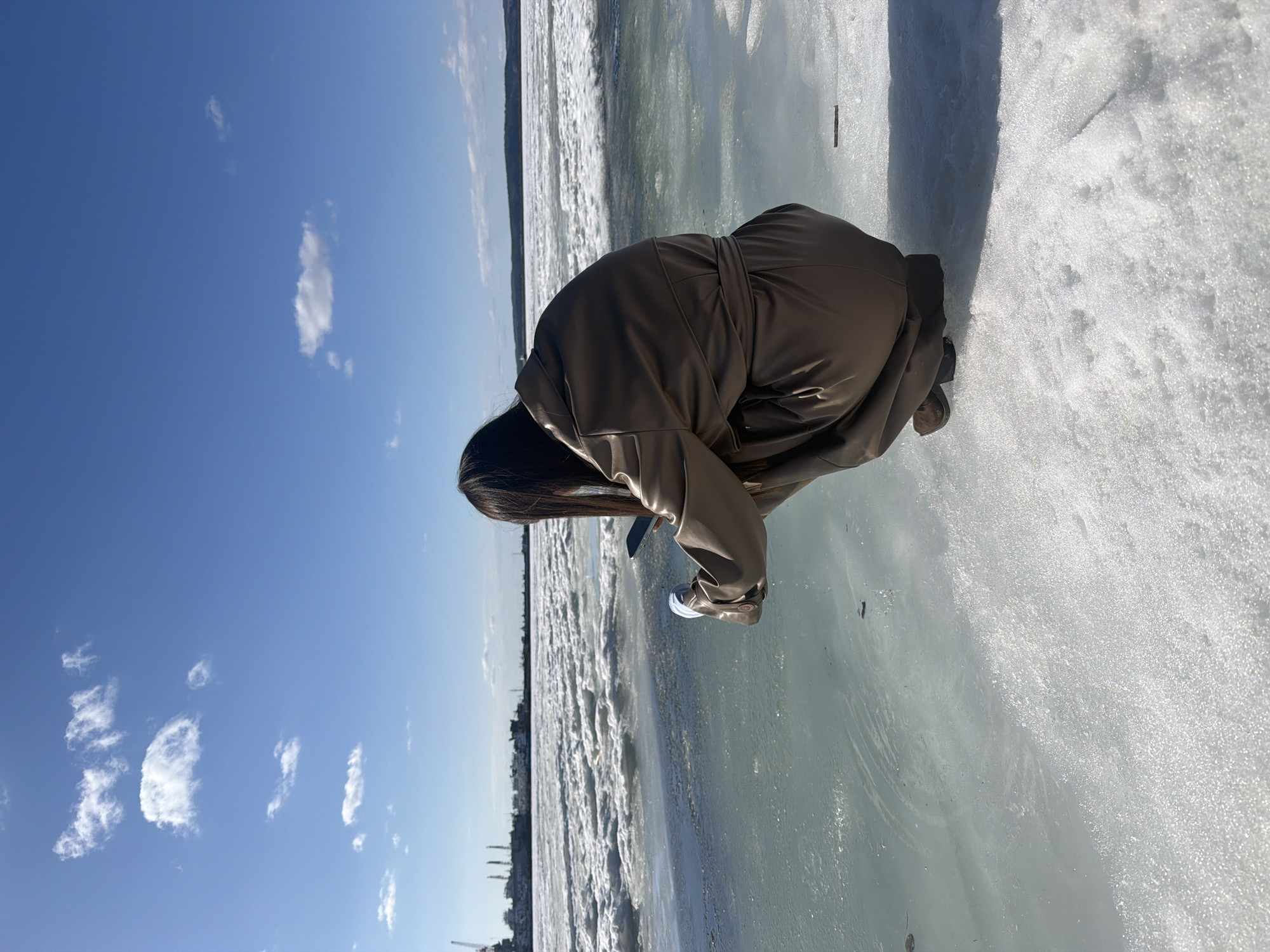 A person is crouching on an icy surface under a clear blue sky with scattered clouds. The person is wearing a brown coat. In the background, there's a cityscape that appears to be Chaikovsky, Russia. The surface beneath the person is a mix of ice and possible frozen water, reflecting the sunlight.