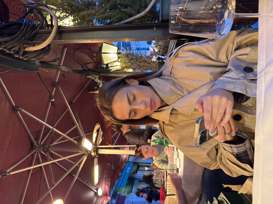 An evening scene depicts a young adult female seated at an outdoor restaurant table in Venice, Italy. The woman, with long dark brown hair and light skin, wears a beige trench coat with visible plaid lining on the rolled cuffs. She is actively engaged with a silver-colored smartphone, held in both hands, looking down at its screen. A silver-toned ring is present on her left ring finger.
The table in the foreground is covered with a white tablecloth. To the right, a large, empty, clear wine glass with a thin stem is positioned. In the midground, other patrons are visible at adjacent tables. To the left, a person with dark blue jeans is partially visible at the same table as the main subject. Further back, another woman with brown hair in a light-colored top is seated, with a green bottle and wine glasses on her table.
Overhead, a large, dark red octagonal umbrella canopy provides cover. Two radiant heaters and several light fixtures are affixed to its frame. A dark vertical pole on the right supports additional outdoor lighting, including a dark lantern with illuminated panels, and features bundles of black cables and a beige hose. A potted evergreen plant is situated to the right of this pole.
The background reveals a narrow urban passage lined with buildings, consistent with Venice's architecture. Blue and white lighting emanates from establishments or structures in the distance, under a dark blue sky, indicating twilight or night.