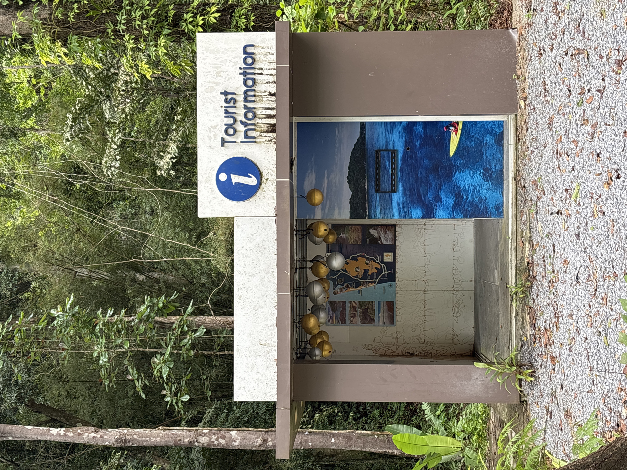 A tourist information kiosk stands in Tambon Thai Mueang, Thailand, surrounded by lush foliage. The kiosk is constructed of brown and white materials with a sign on top that reads "Tourist Information" next to an "i" symbol. Inside the kiosk are various informational displays, including images of a beach scene with a person on a yellow kayak, a map of the area, and other related pictures. A string of gold and silver spheres hangs within the kiosk. The ground around the kiosk is covered in small gravel and foliage. Trees and dense greenery fill the background.