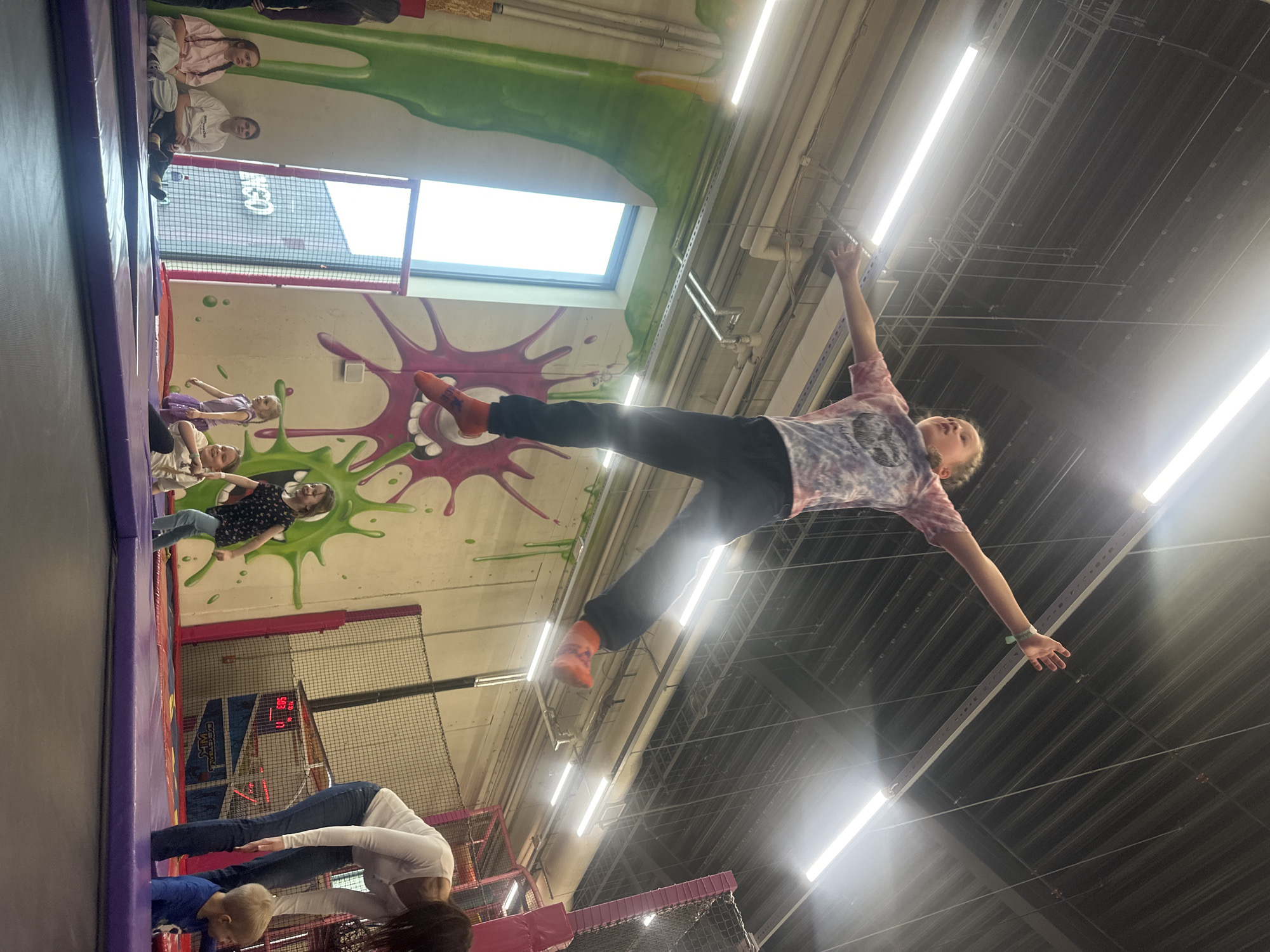 The image shows a child mid-air, apparently jumping or being propelled upwards inside an indoor trampoline park. The child, wearing a tie-dye shirt, dark pants, and orange socks, has their arms outstretched. The trampoline park features brightly colored walls with splattered paint designs. Other people are visible in the background, standing or sitting near the trampolines. Skylights provide natural light, and fluorescent lighting fixtures are also visible on the ceiling. The image was taken in Riga, Latvia.