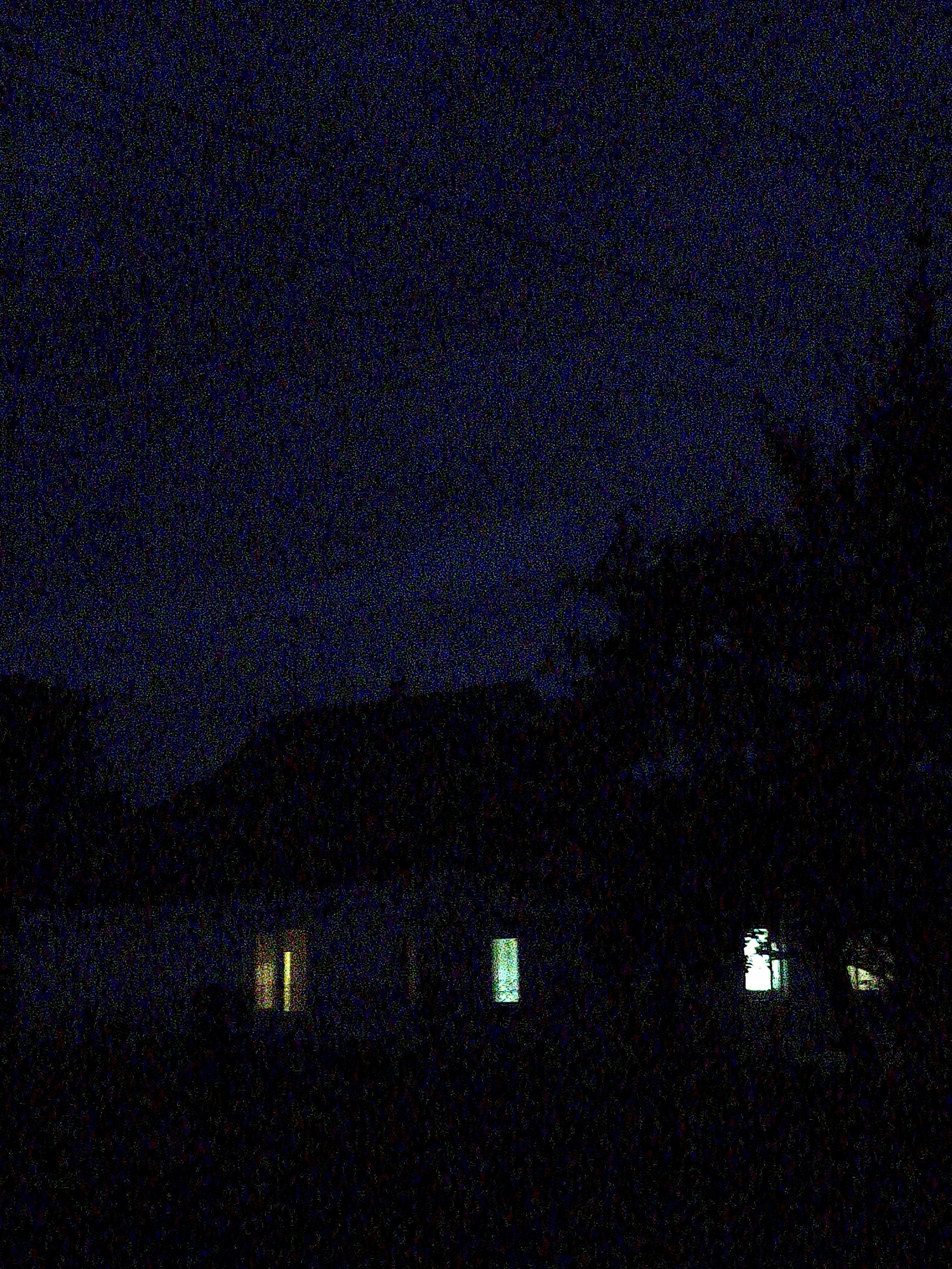 This image depicts a nighttime scene in Ustyvytsia, Ukraine, characterized by extremely low light and significant digital noise across the entire frame. The upper portion of the image shows a dark blue, heavily pixelated sky. A dark, indistinct silhouette of what appears to be a distant landscape or structures forms a horizon line across the mid-ground.
In the foreground, a dark building, likely a house, is largely obscured by shadows and noise. Several windows on this structure are illuminated:
* On the left, a window displays two distinct vertical yellowish-orange lights.
* Towards the center-left, another window emits a rectangular greenish-blue light.
* Further to the right, two more windows are brightly lit with white/yellow light, revealing some internal structure or window frames.
To the far right of the building, a large mass of dark, indistinct foliage or trees is visible, heavily silhouetted against the slightly lighter sky. No individuals, specific actions, or interactions are discernible within the scene.