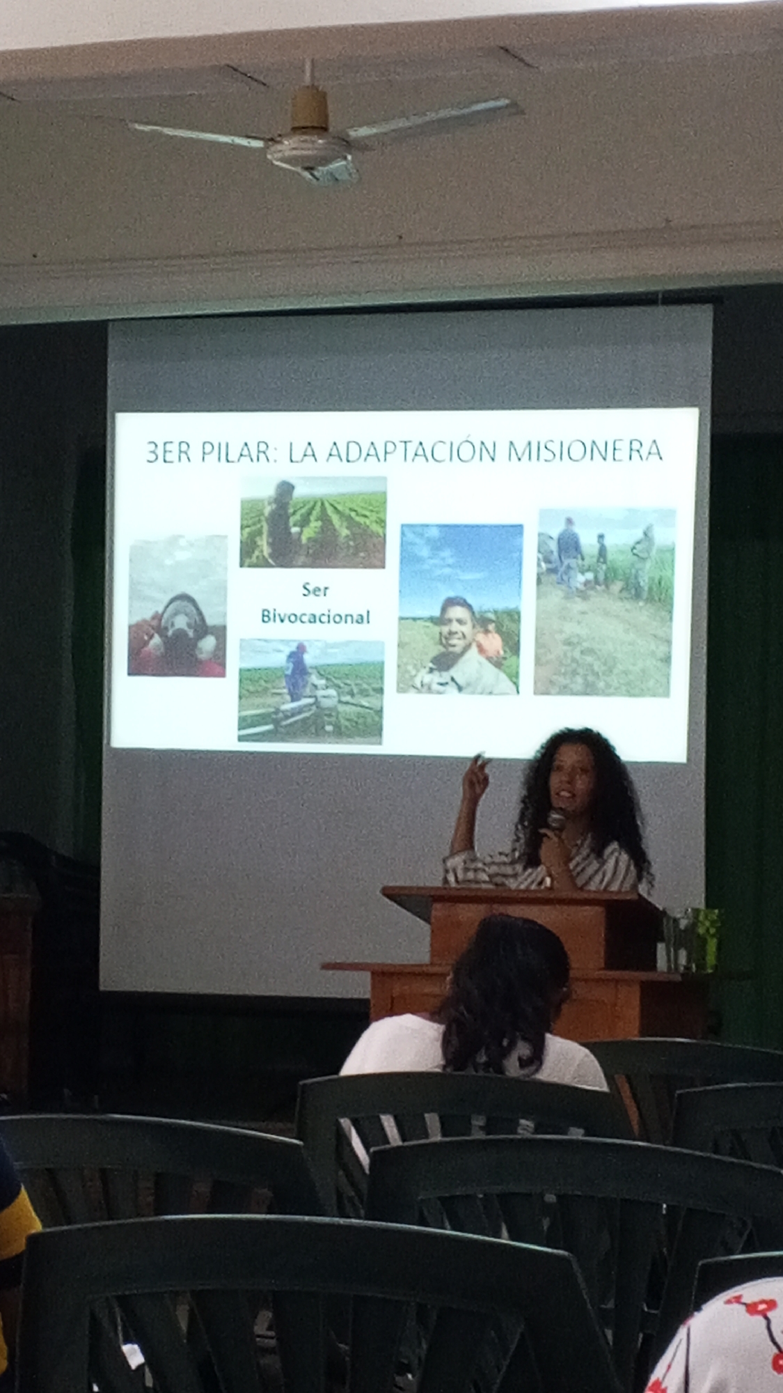 A woman with curly dark hair stands at a wooden podium, speaking into a microphone, in front of a projection screen displaying the text "3ER PILAR: LA ADAPTACIÓN MISIONERA" and several smaller images. A person with dark hair wearing a white shirt sits in a chair in the foreground, facing away from the camera. The room is filled with green plastic chairs. A ceiling fan is visible in the upper part of the frame. The location is Unknown, Unknown.