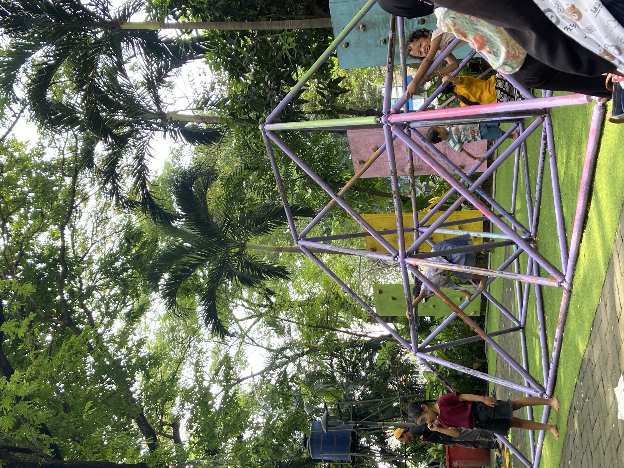 The image shows a colorful geodesic dome climbing structure in a park, likely in Surabaya, Indonesia. Children are actively engaged in climbing on the dome, while other children are standing near the base. Palm trees and other greenery surround the play area. The ground around the dome is covered in artificial turf and brick pathways.