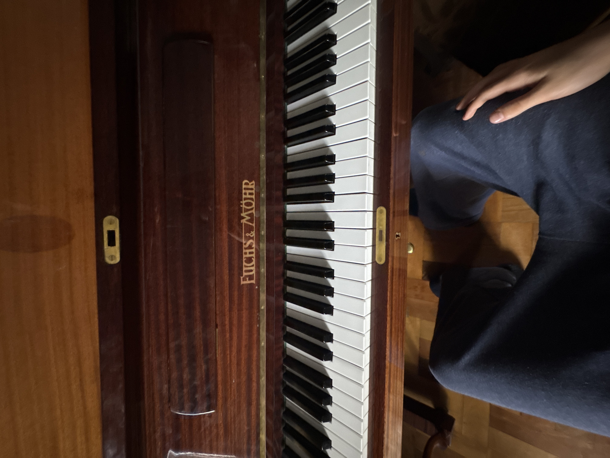 The image provides an overhead view of a person seated at a dark wooden piano in an indoor setting, identified as Yerevan, Armenia. The piano's keyboard, featuring white and black keys, extends down the center of the frame. To the left of the keys, the brand name "FUCHS & MÖHR" is clearly visible in gold lettering on the polished, dark wood surface, which displays a fine grain. Small, rectangular gold-colored metal plates, possibly decorative or part of a locking mechanism, are present on the far left and right edges of the piano's wooden frame. A person's bare hand rests gently on their leg, clad in dark blue fabric (likely trousers or sweatpants), positioned to the right of the keyboard. Parts of a light-colored wooden floor are visible beneath and around the piano. The subdued lighting suggests an indoor scene, likely during the evening or night. The image captures a quiet moment at the instrument, either in anticipation of playing or in a pause from it.