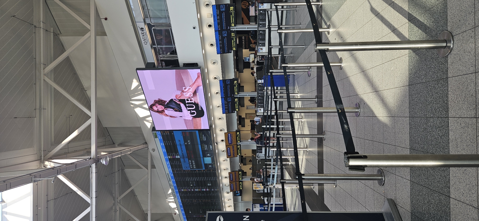 The image shows the departures hall of Budapest Ferenc Liszt International Airport. In the background, a large digital screen displays an advertisement for GUESS featuring Chiara Ferragni. To the left of the screen, a flight information board lists departures. In the foreground, queuing lanes with stanchions and black belts guide passengers towards check-in counters. Several check-in counters are visible, with some Ryanair signage. Passengers are seen at the counters and walking through the hall. The environment is bright, with sunlight casting shadows across the tiled floor.